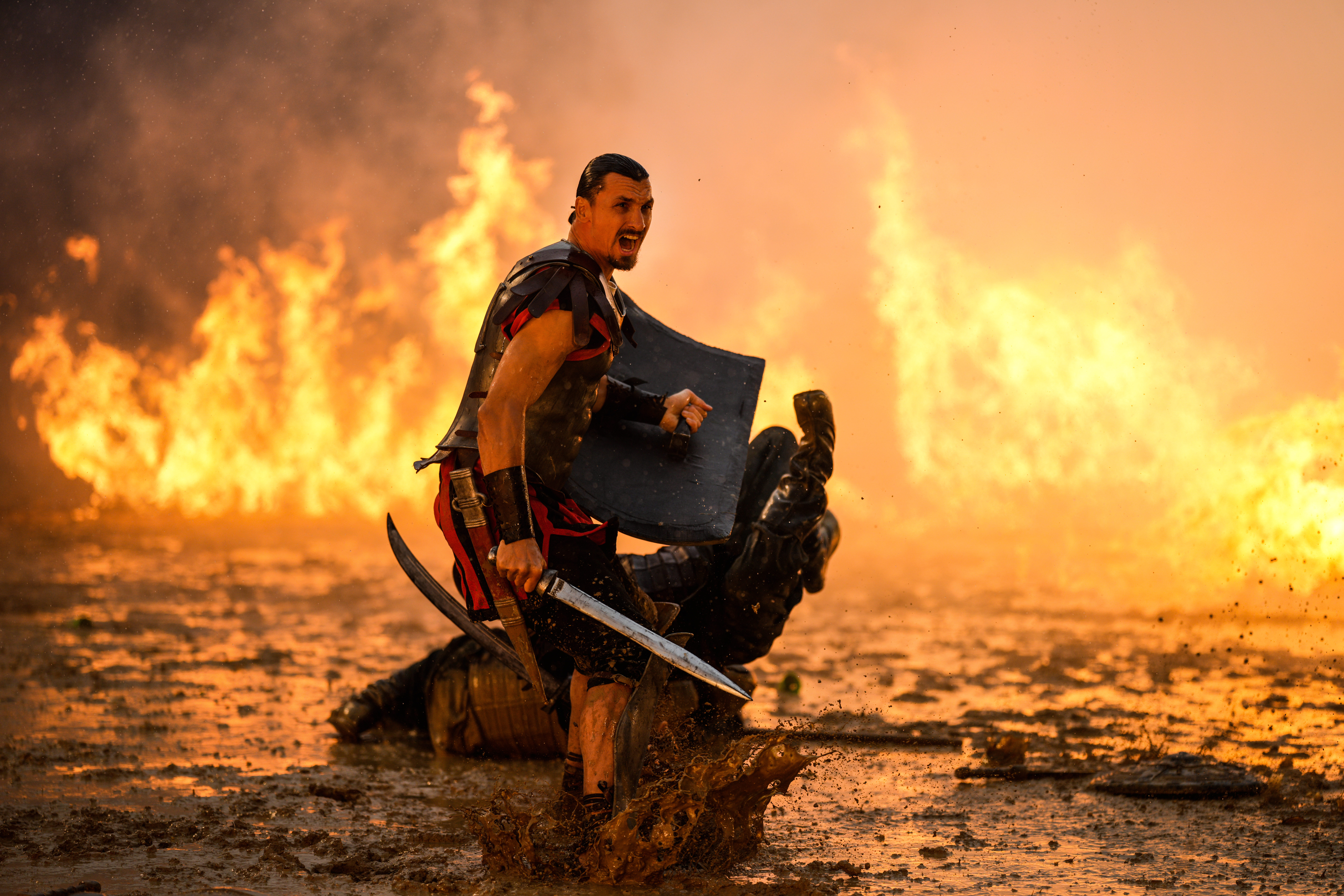 Ein Gladiator in Rüstung mit Schwert steht über einem gefallenen Gegner in feuriger Umgebung.