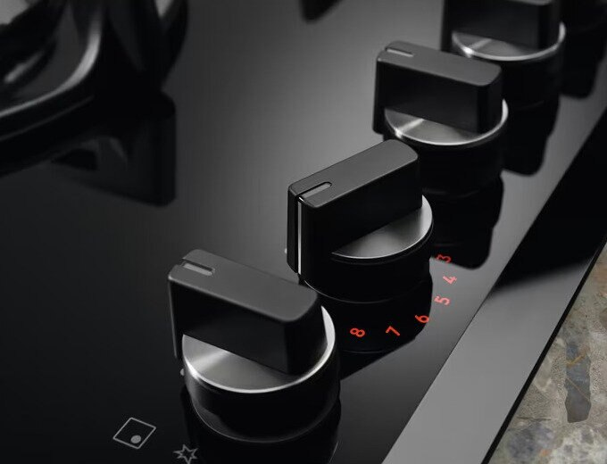 Zbliżenie czarnych pokręteł na szklanej płycie grzejnej z czerwonymi oznaczeniami numerycznymi.