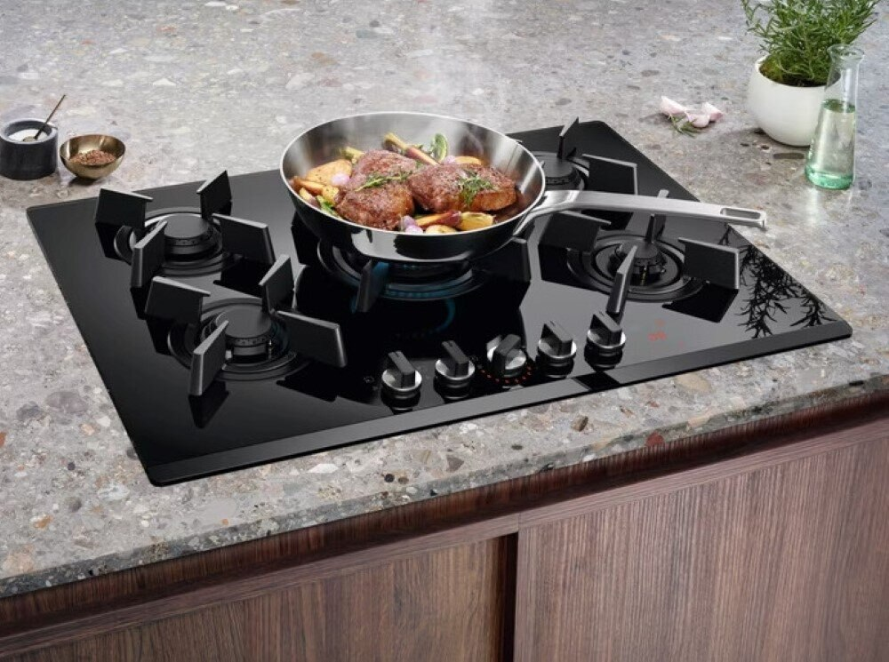 Czarna płyta gazowa z patelnią i dwoma garnkami. Jedzenie gotuje się na patelni. Tło to blat kuchenny.