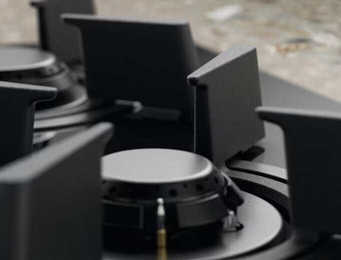 Zbliżenie palnika gazowego na kuchence. Pierwszy plan: czarny palnik i otaczające elementy. Tło: rozmyta kuchenka.