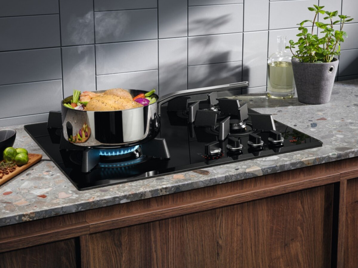 Płyta grzewcza z garnkiem jedzenia gotującego się na niebieskim płomieniu. Widoczne warzywa i mięso. Blat z marmuru i drewna.