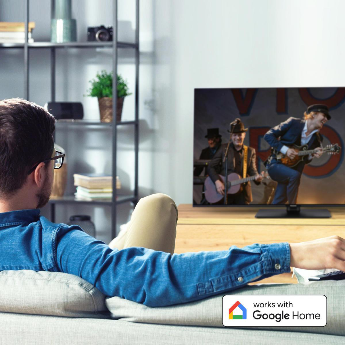 Mann schaut TV, Szene mit Band auf dem TV. Sofa, Regal und Google Home Logo sichtbar.