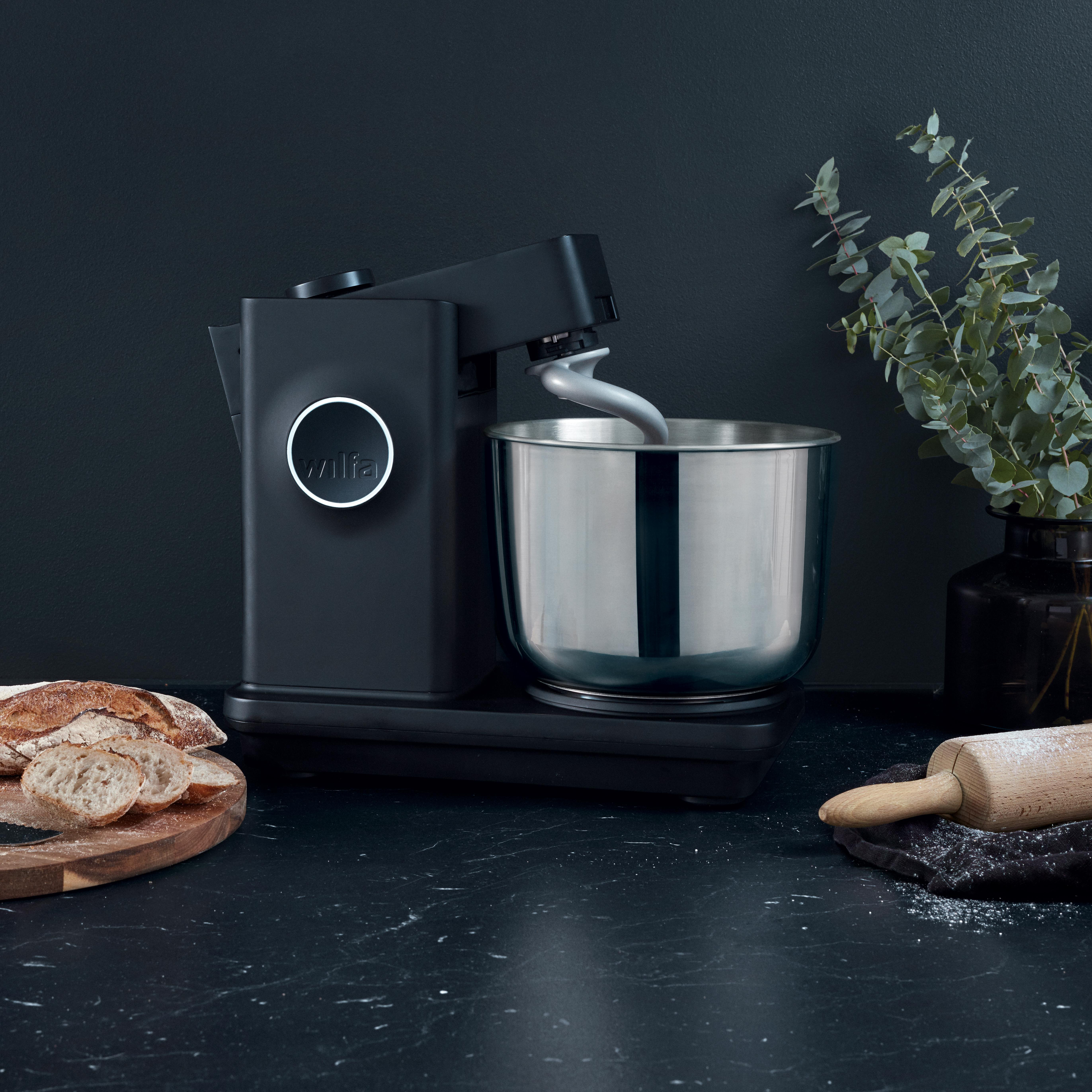 Schwarze Küchenmaschine mit Brot und Nudelholz auf dunkler Oberfläche. Pflanze in Vase.