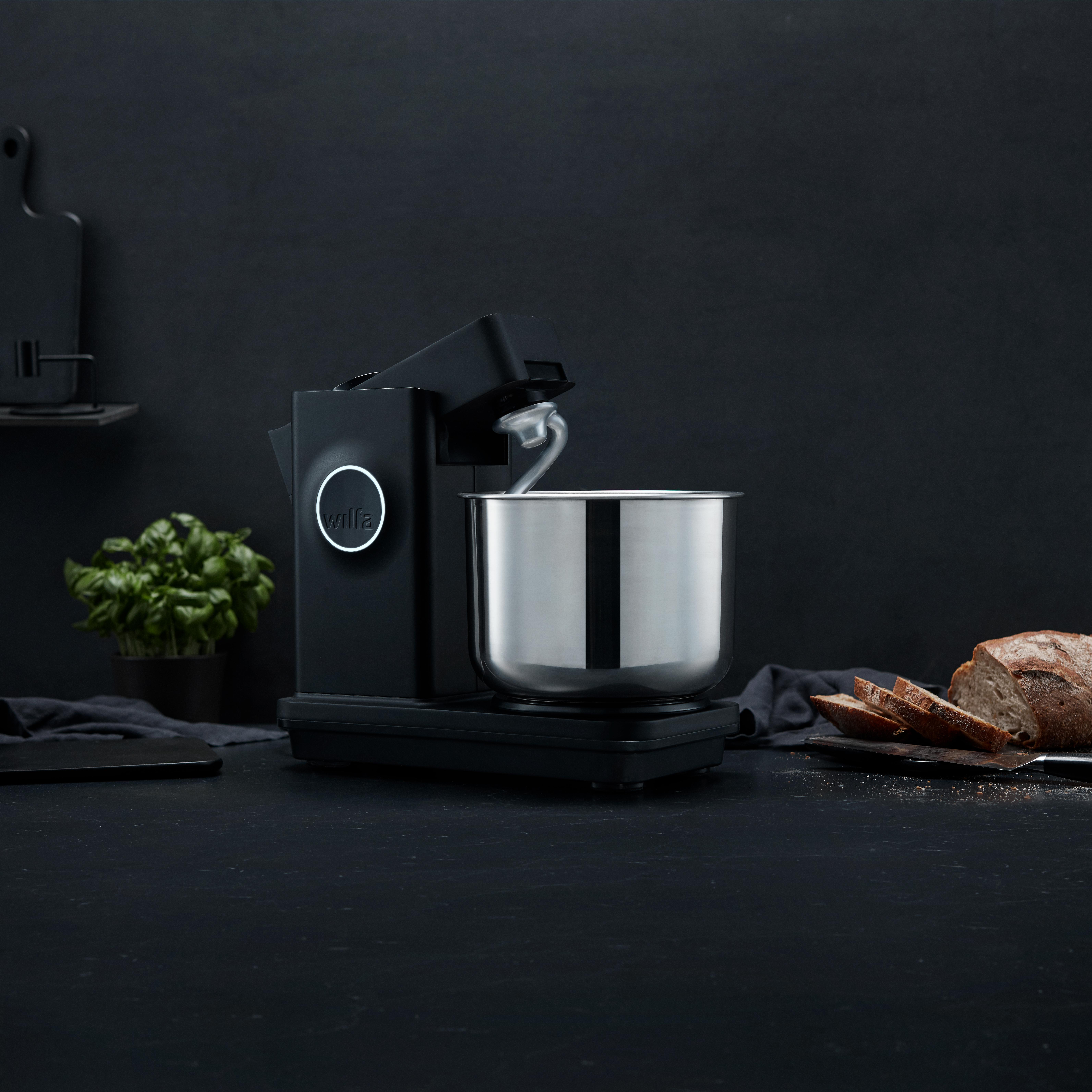 Schwarzer Mixer mit Edelstahlschüssel auf dunkler Oberfläche mit kleiner Pflanze und Brot. Dunkler Hintergrund.