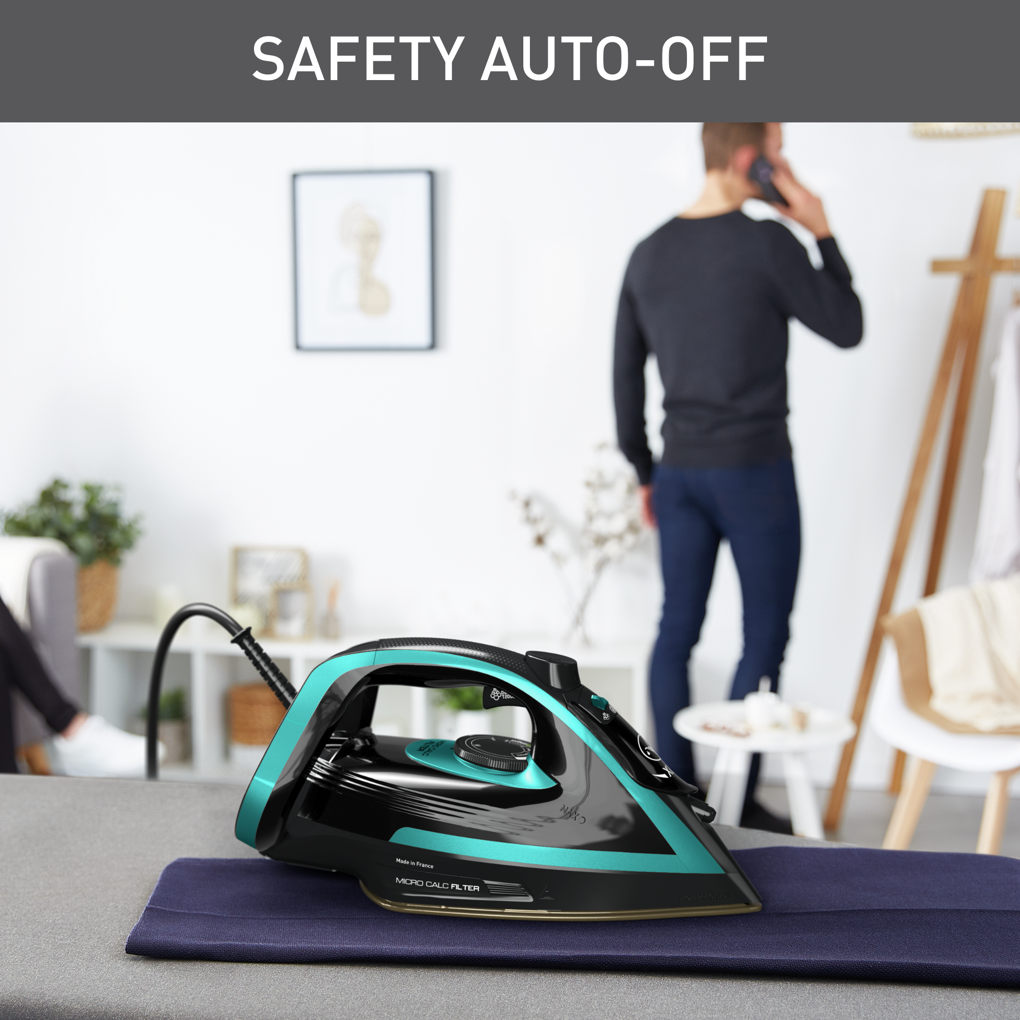 Egy fekete és türkizkék díszítésű vasaló egy deszkán, egy férfi telefonál a háttérben. A SAFETY AUTO-OFF felirat.