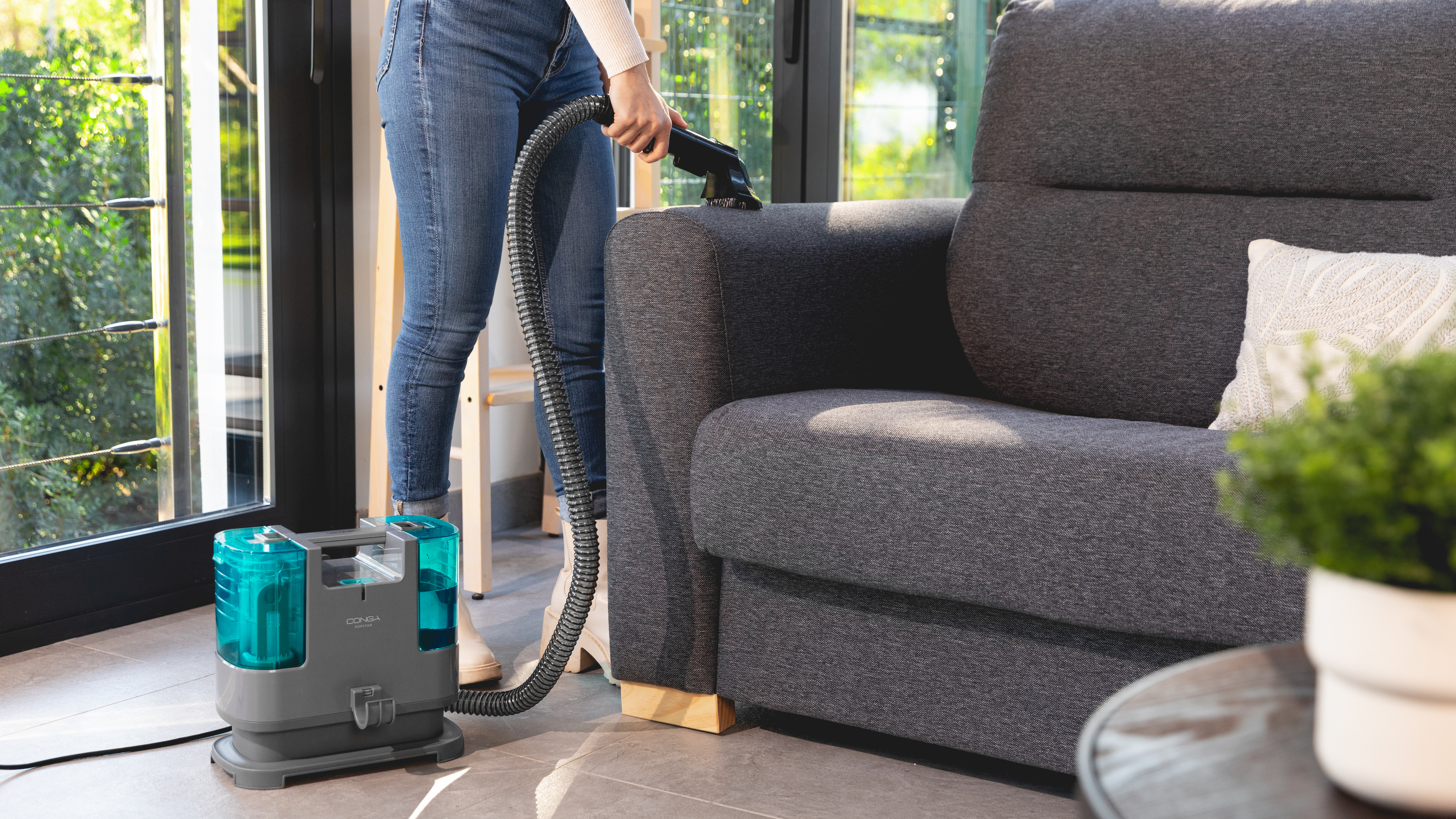 Una persona aspira un sofá gris con una máquina de limpieza. La máquina es turquesa y gris.