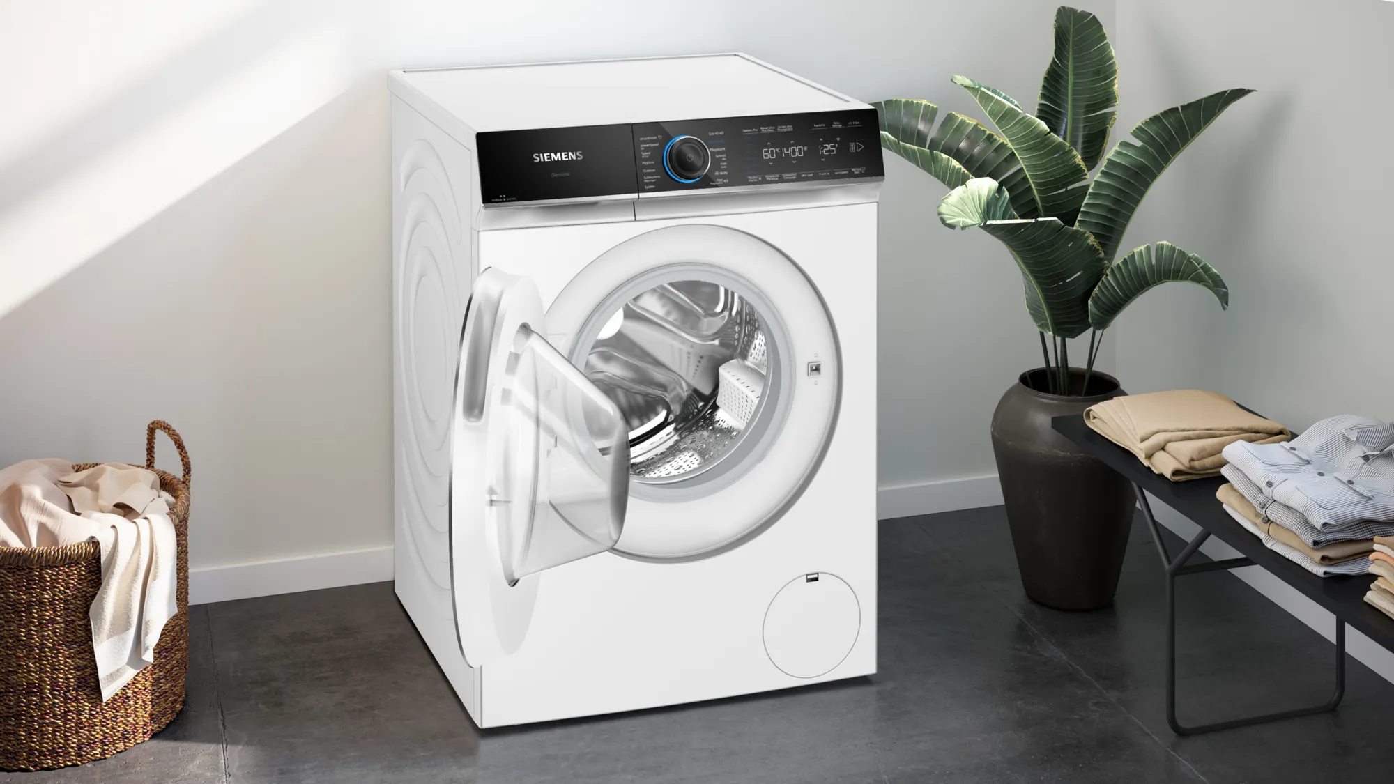 Weiße Waschmaschine mit offener Tür. Wäschekorb und Pflanze im Hintergrund.