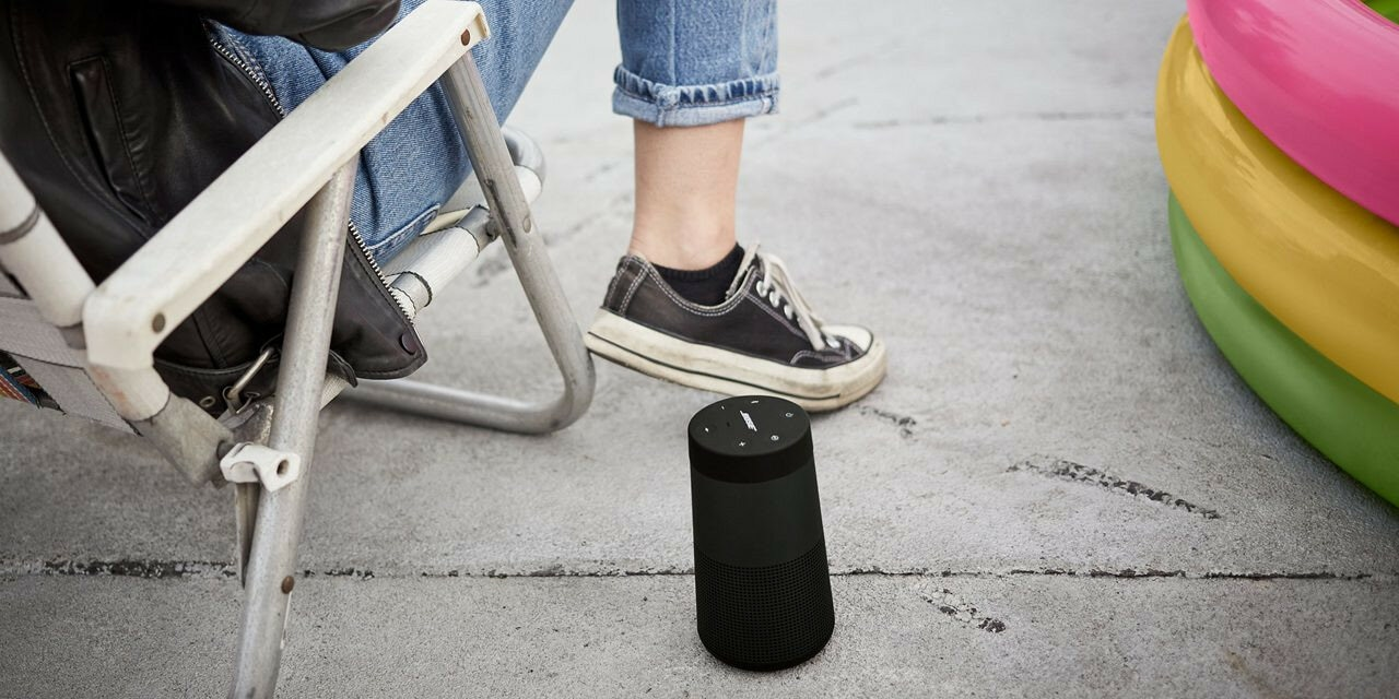 Osoba siedząca na krześle z czarnym głośnikiem w pobliżu. Tło obejmuje betonową podłogę i dmuchane zabawki do basenu.