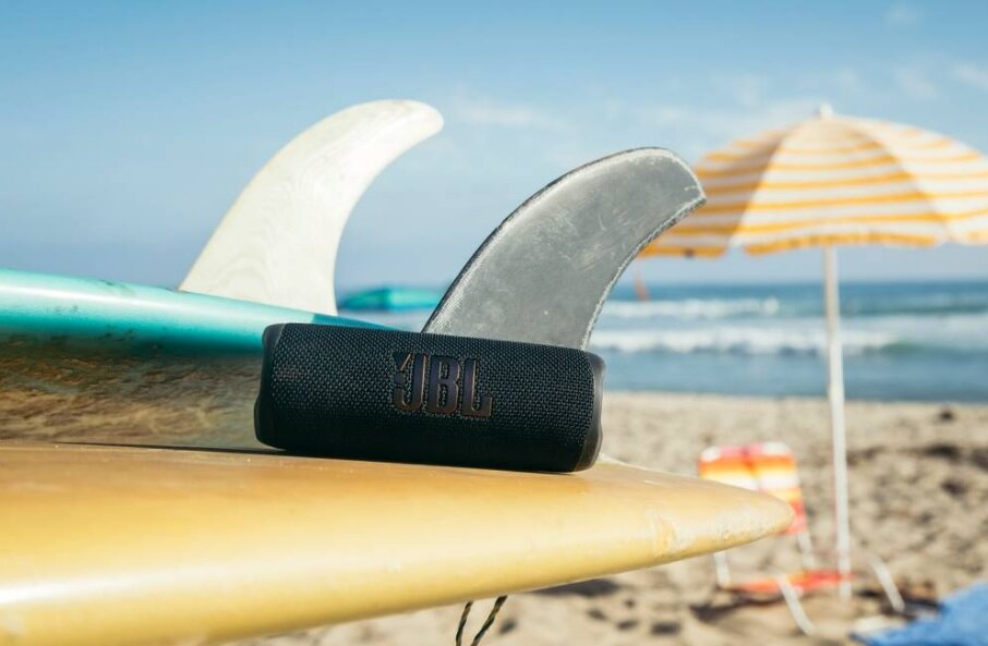 Czarny głośnik JBL na desce surfingowej. Deski surfingowe, plaża i parasol w tle.