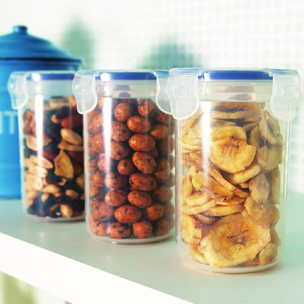 Trzy przezroczyste słoiki z niebieskimi pokrywkami. Każdy słoik zawiera inne produkty spożywcze: orzechy, orzeszki ziemne w polewie i chipsy bananowe.