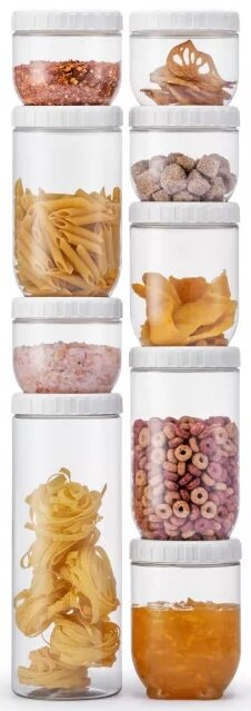 Szklane słoiki wypełnione produktami spożywczymi są ułożone w stos. Zawartość obejmuje makaron, płatki, przyprawy i inne produkty.