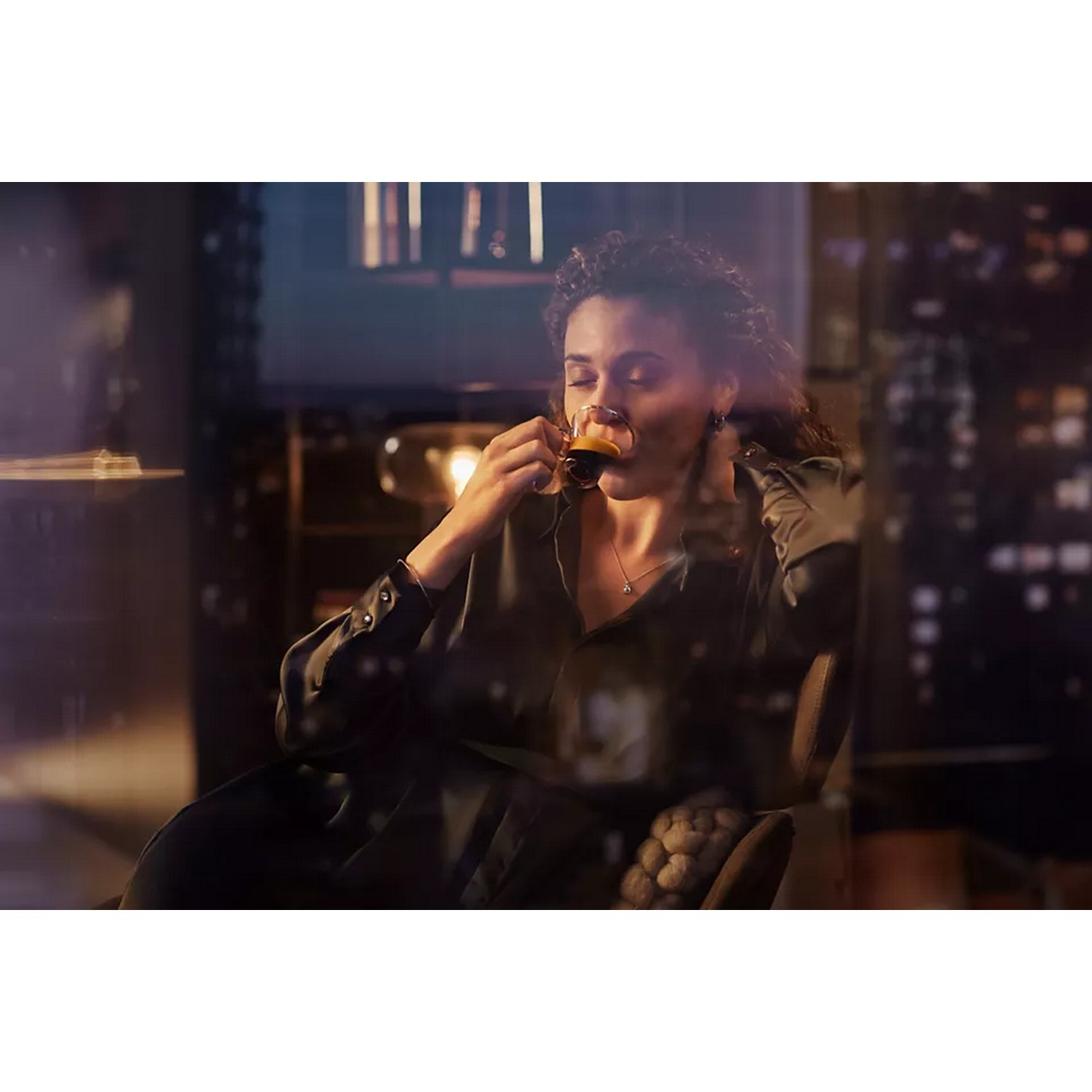 Woman drinking coffee in a dimly lit room. City lights outside window.