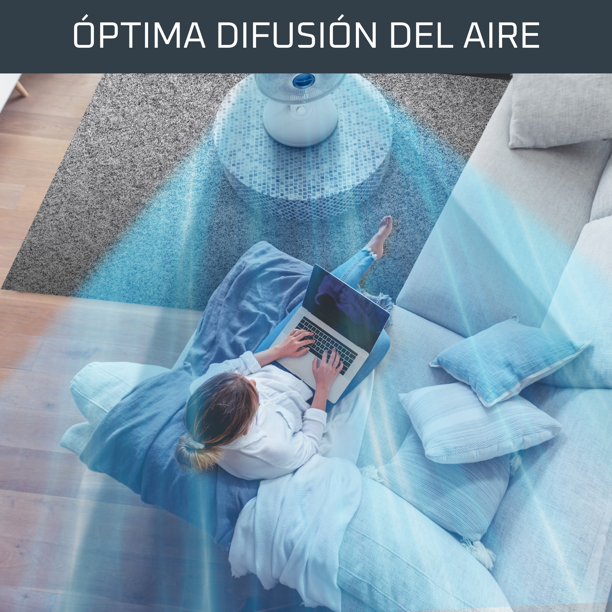 Mujer sentada en un sofá con portátil, con un ventilador al fondo.