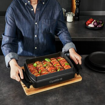 Osoba trzymająca tacę z ugotowanym jedzeniem na drewnianej desce do krojenia.