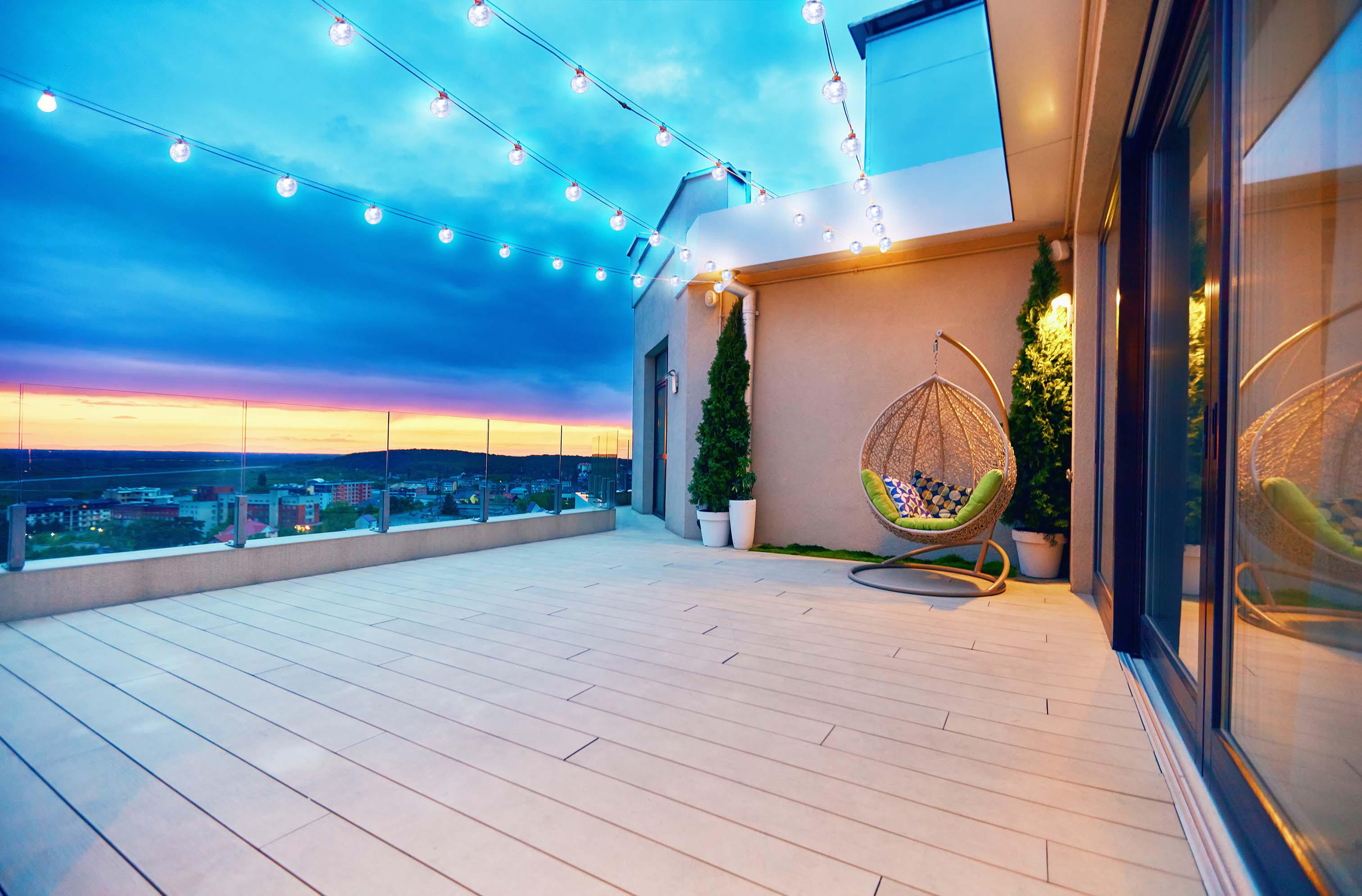 Dachterrasse in der Abenddämmerung mit Lichterketten. Hängestuhl, Bäume und Stadtblick.