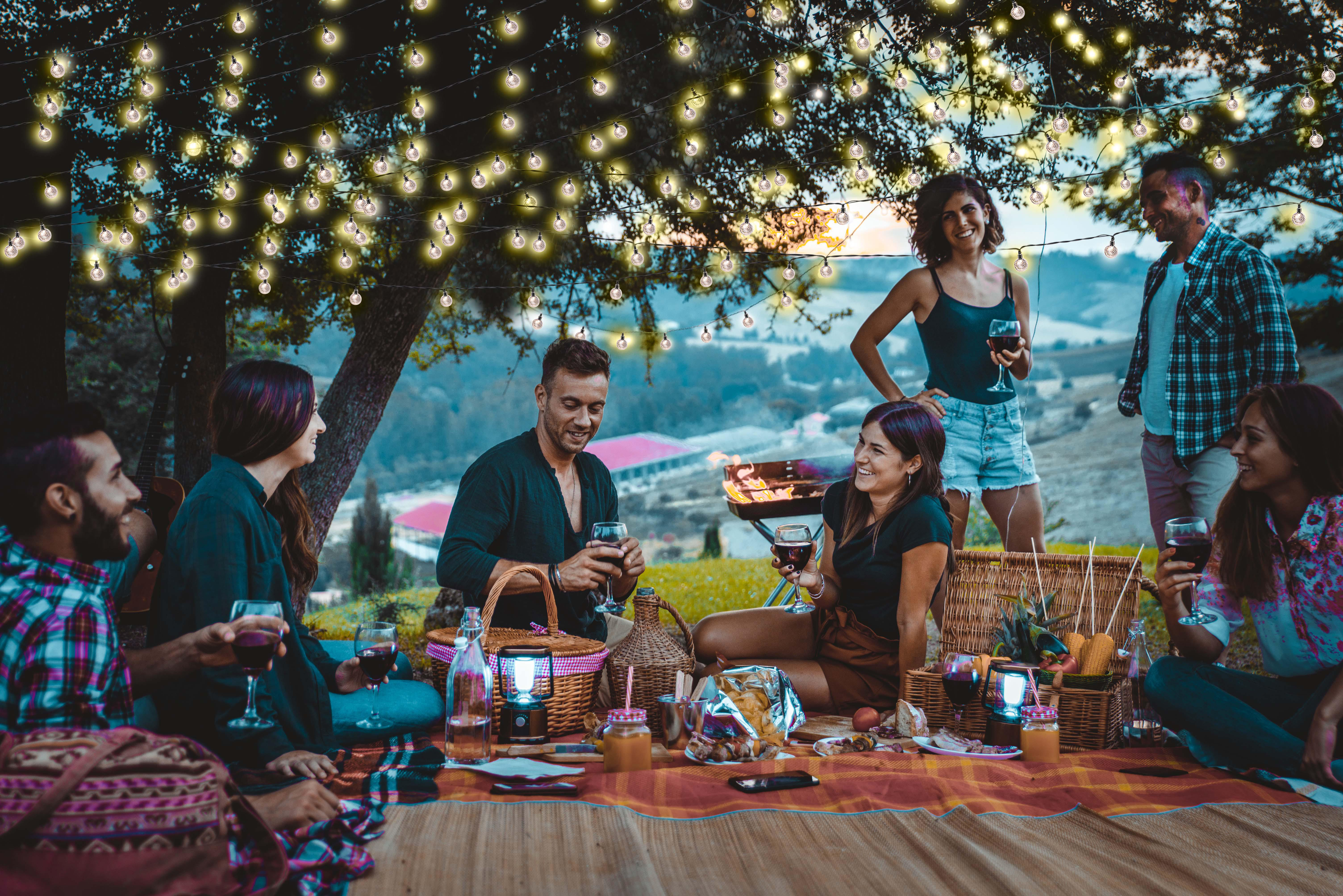 Freunde picknicken im Freien mit Essen, Getränken und Lichterketten.