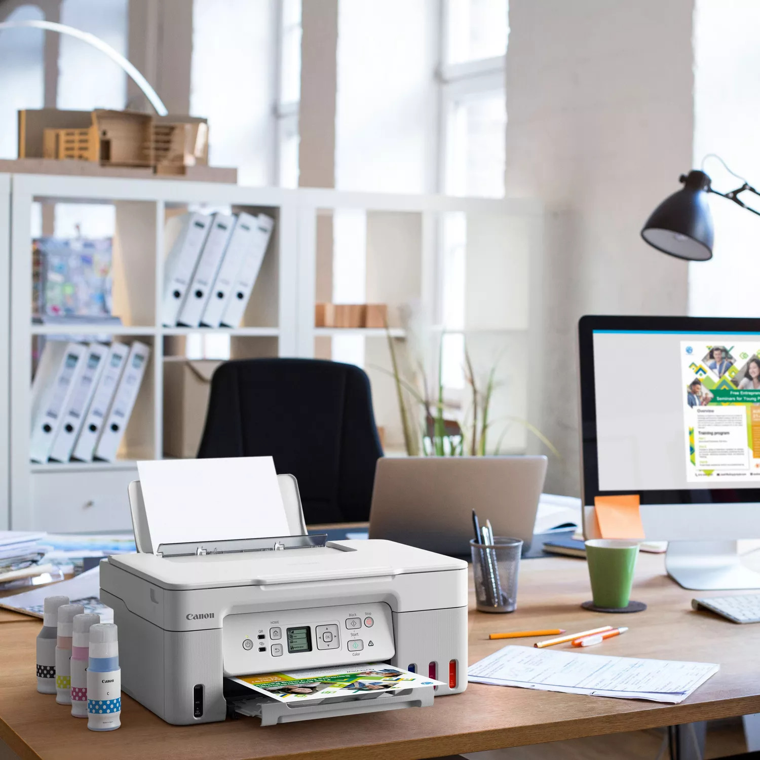 Witte Canon-printer op een houten bureau. Inktflessen, een computer en kantoorbenodigdheden in de buurt.