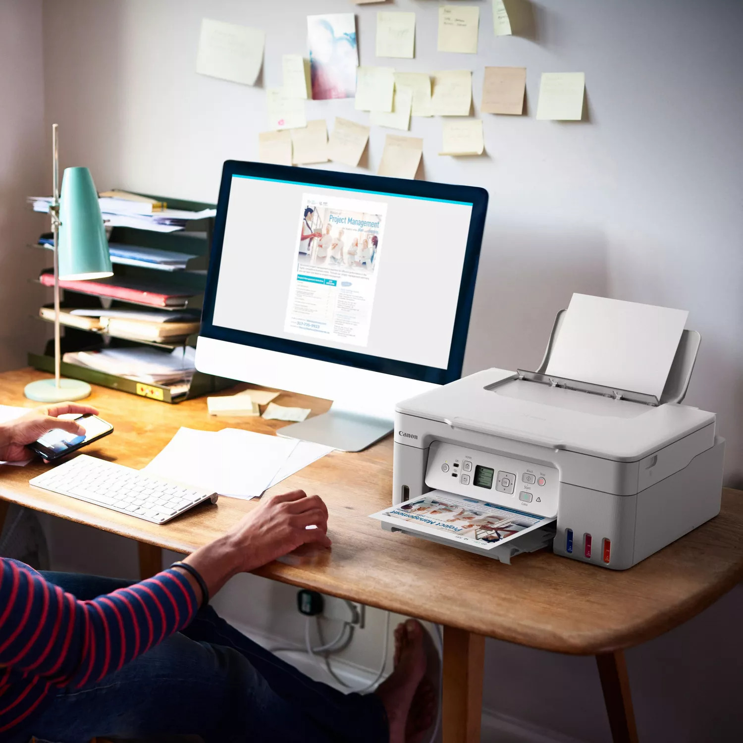 Een persoon zit aan een bureau met een computer waarop een document wordt weergegeven. Een printer drukt hetzelfde document af. Er zitten plakbriefjes op de muur.