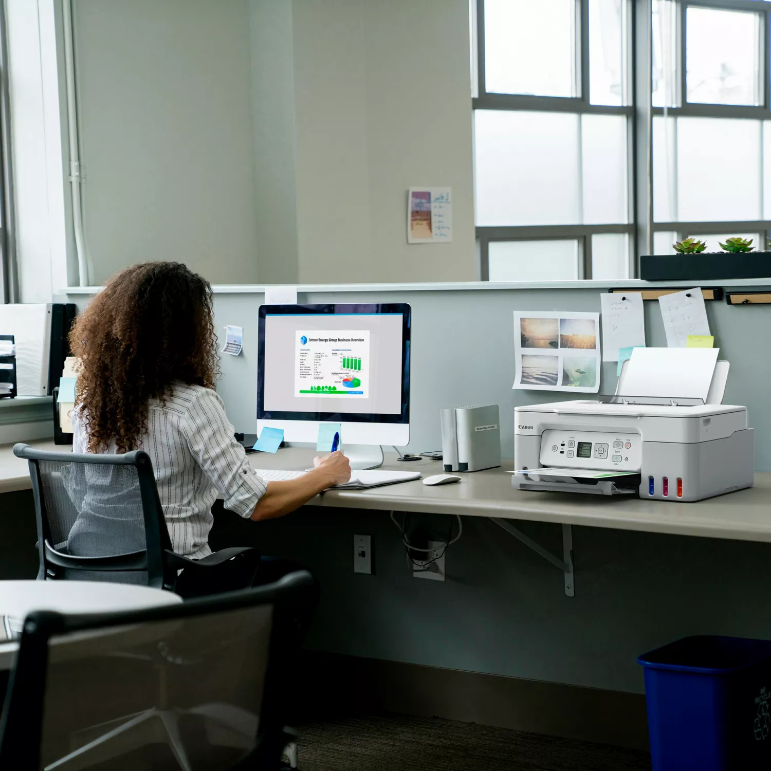Een vrouw met krullend haar zit aan een bureau en werkt op een computer met een zakelijk overzicht. Een printer en een prullenbak staan in de buurt.