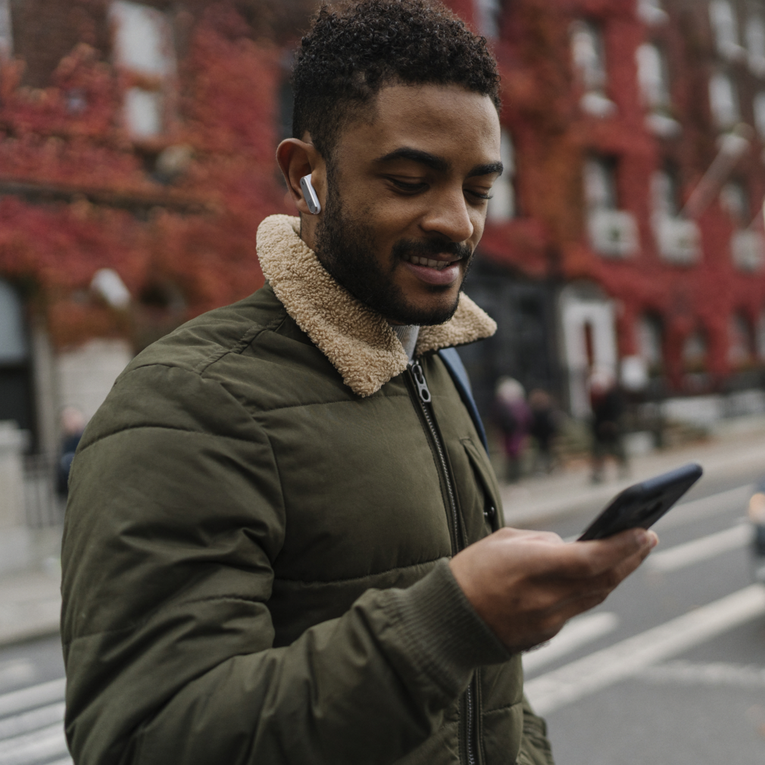 A man with earbuds and a phone, walking on a city street with red ivy.