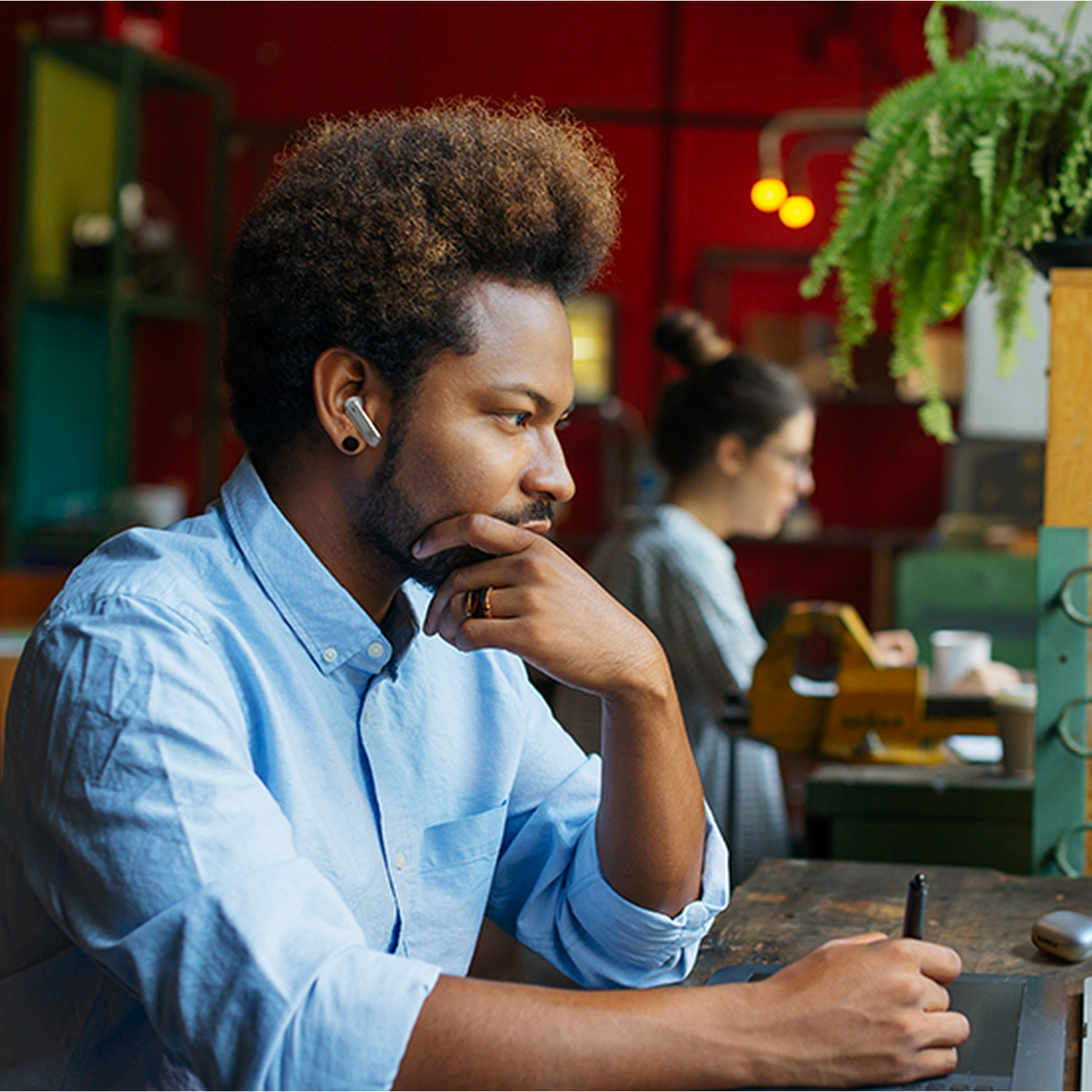 A man with earbuds in an office setting, looking thoughtfully while working on a tablet.
