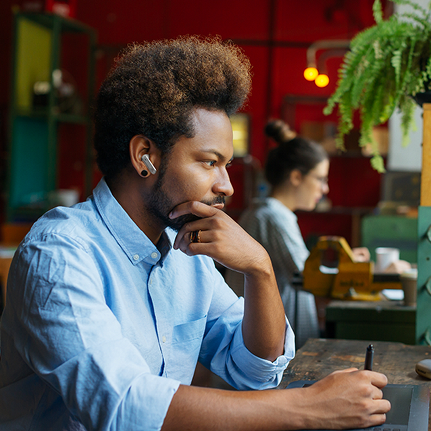Man with earbuds, looking at a screen, holding a stylus. A woman works in background.