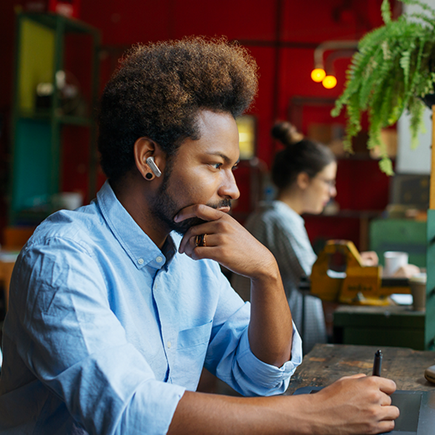 A man in a blue shirt wearing earbuds, working at a table. A woman is in the background.