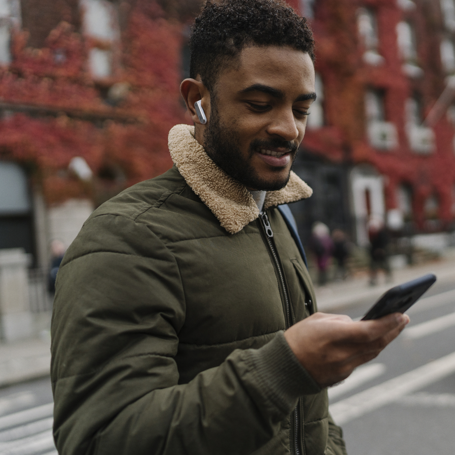 Man in jacket with earbuds, holding phone, smiling. Buildings with red vines in the background.