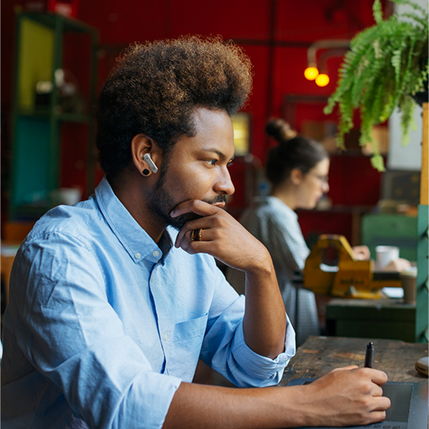 Man with earbuds, looking at a screen, holding a stylus. A woman works in background.