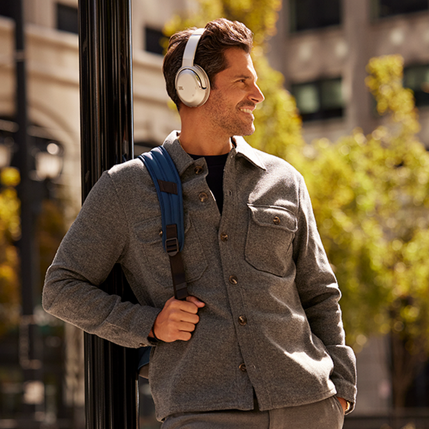 Man wearing white headphones and a blue backpack leaning against a pole, smiling.