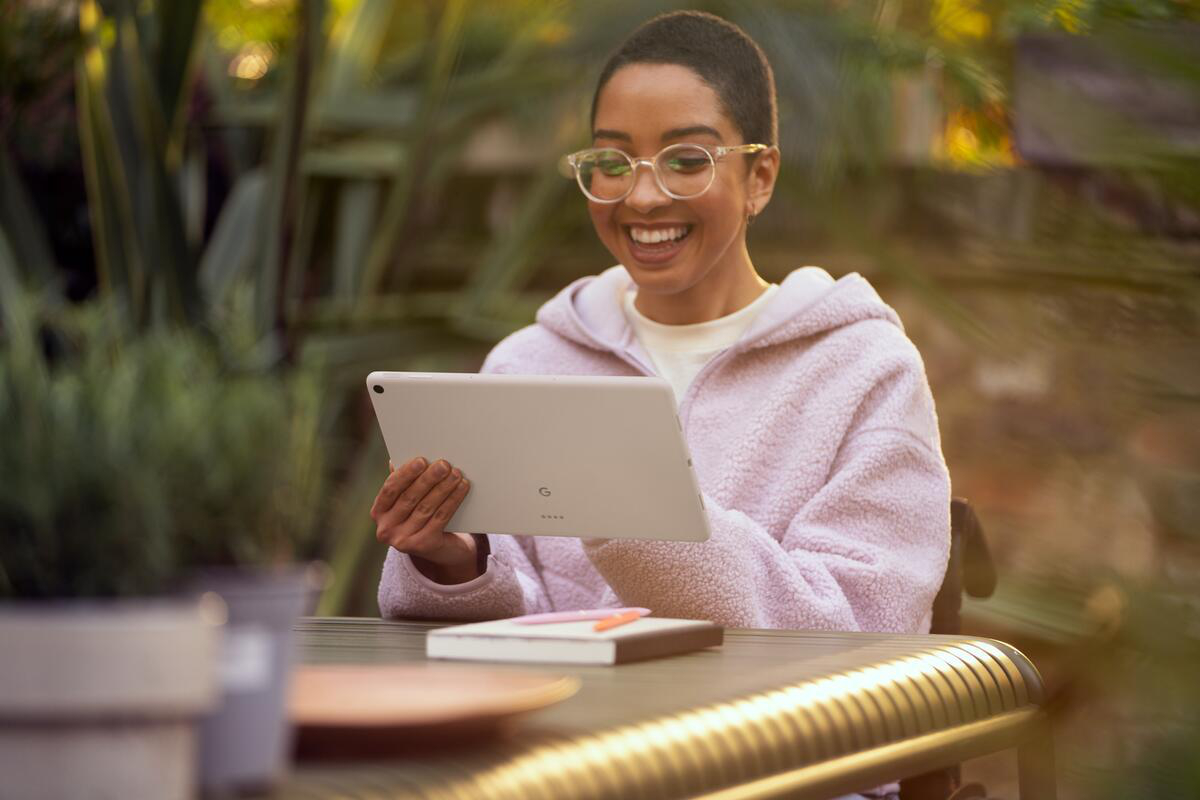 Eine Frau mit Brille lächelt, während sie draußen auf ein Tablet schaut. Pflanzen sind im Hintergrund.