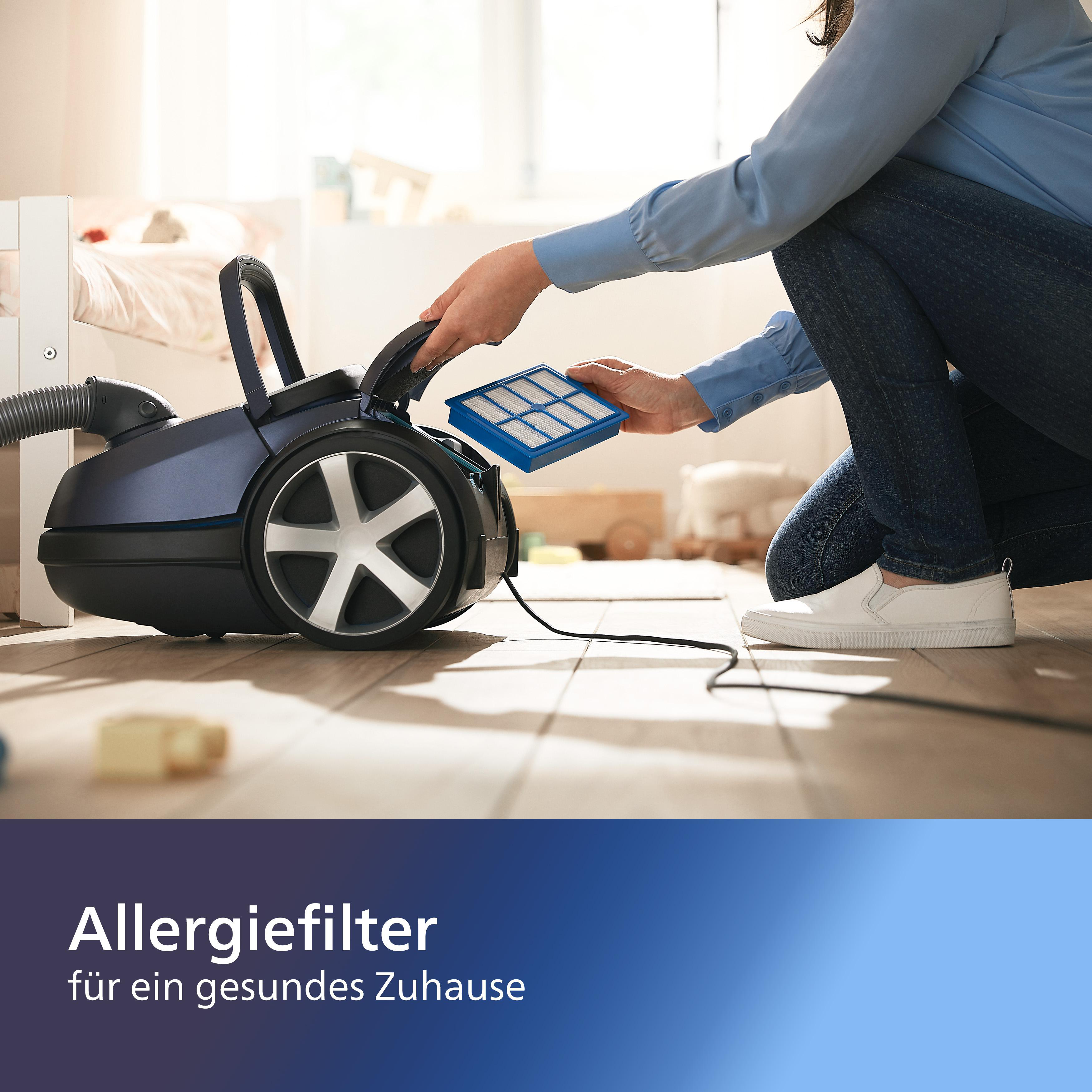 Person tauscht blauen Filter in Staubsauger. Der Staubsauger ist schwarz und silber. Im Hintergrund ist ein Bett.
