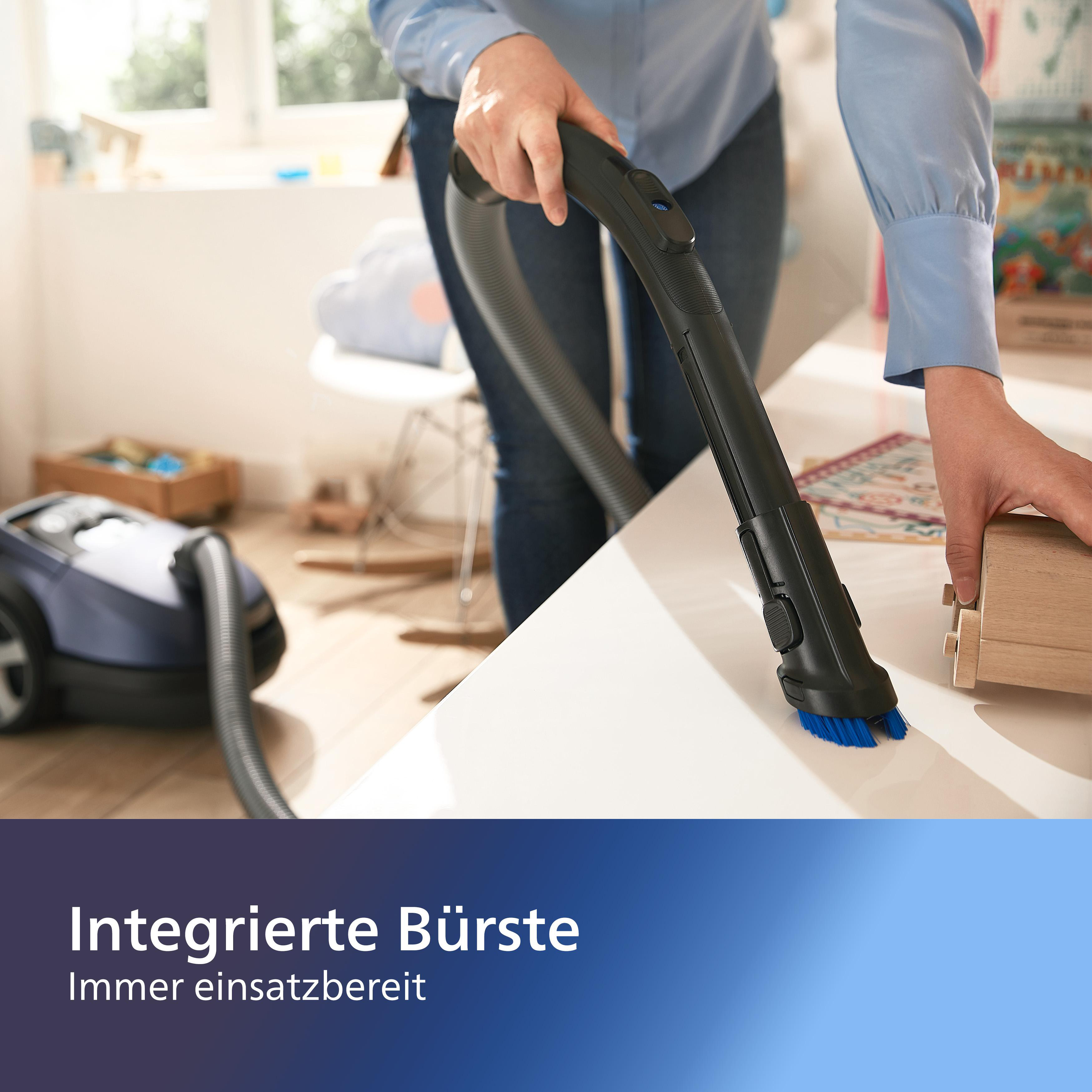 Person verwendet Staubsauger mit blauer Bürste. Maschine steht auf weißem Tisch in einem Raum.
