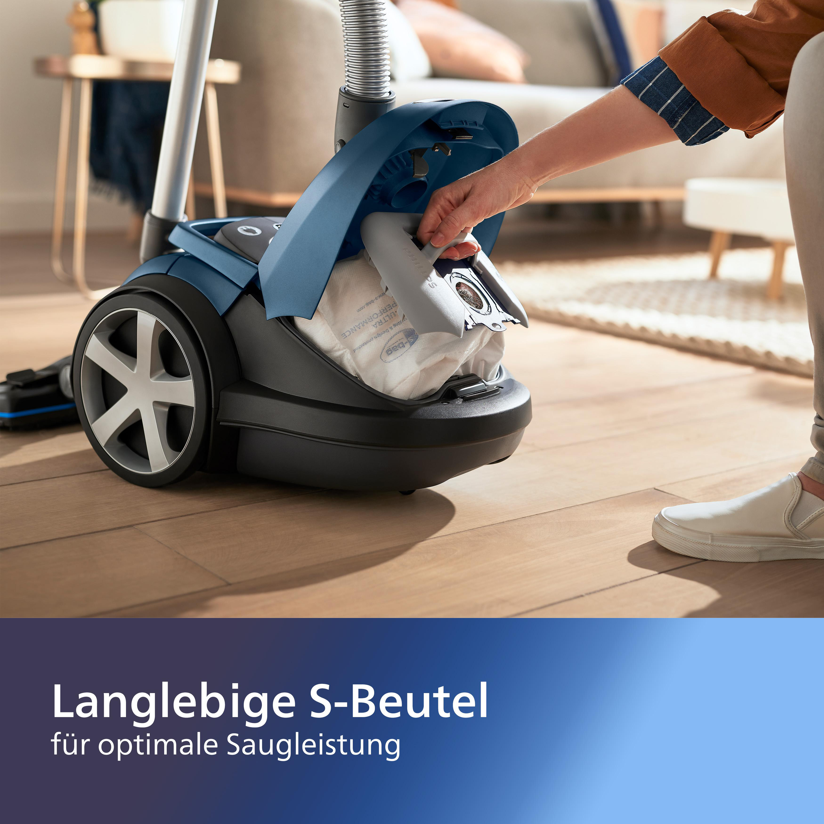 Eine Person entnimmt einen Staubbeutel aus einem blau-schwarzen Staubsauger auf einem Holzboden. Im Hintergrund ein Wohnzimmer.