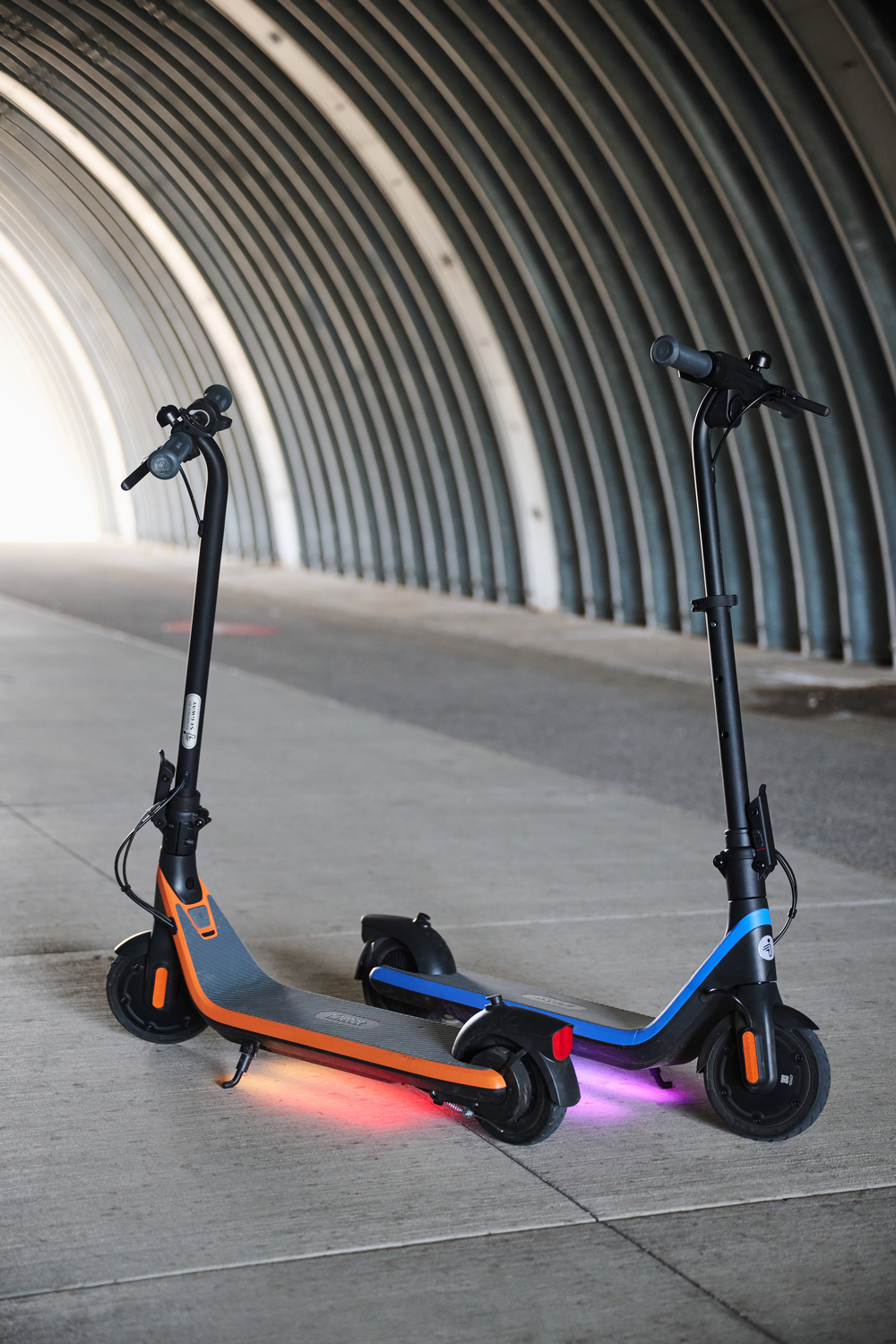 Dos patinetes, naranja y azul, estacionados en un paso subterráneo con luces encendidas.