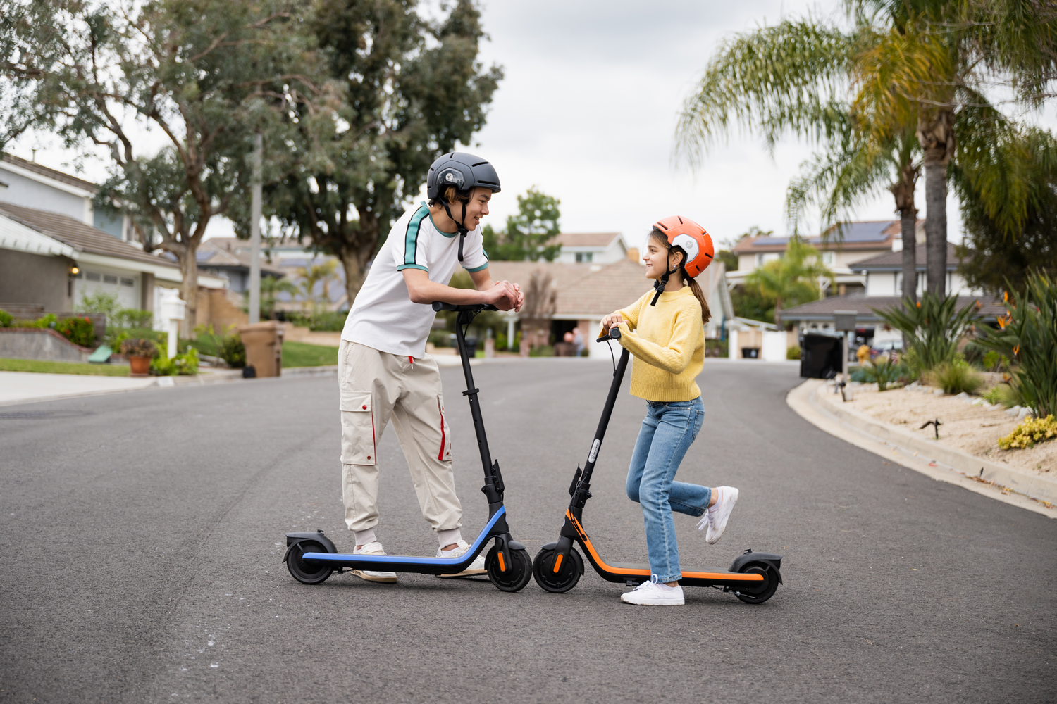 Dos niños andan en patinetes en una calle residencial. Llevan cascos.