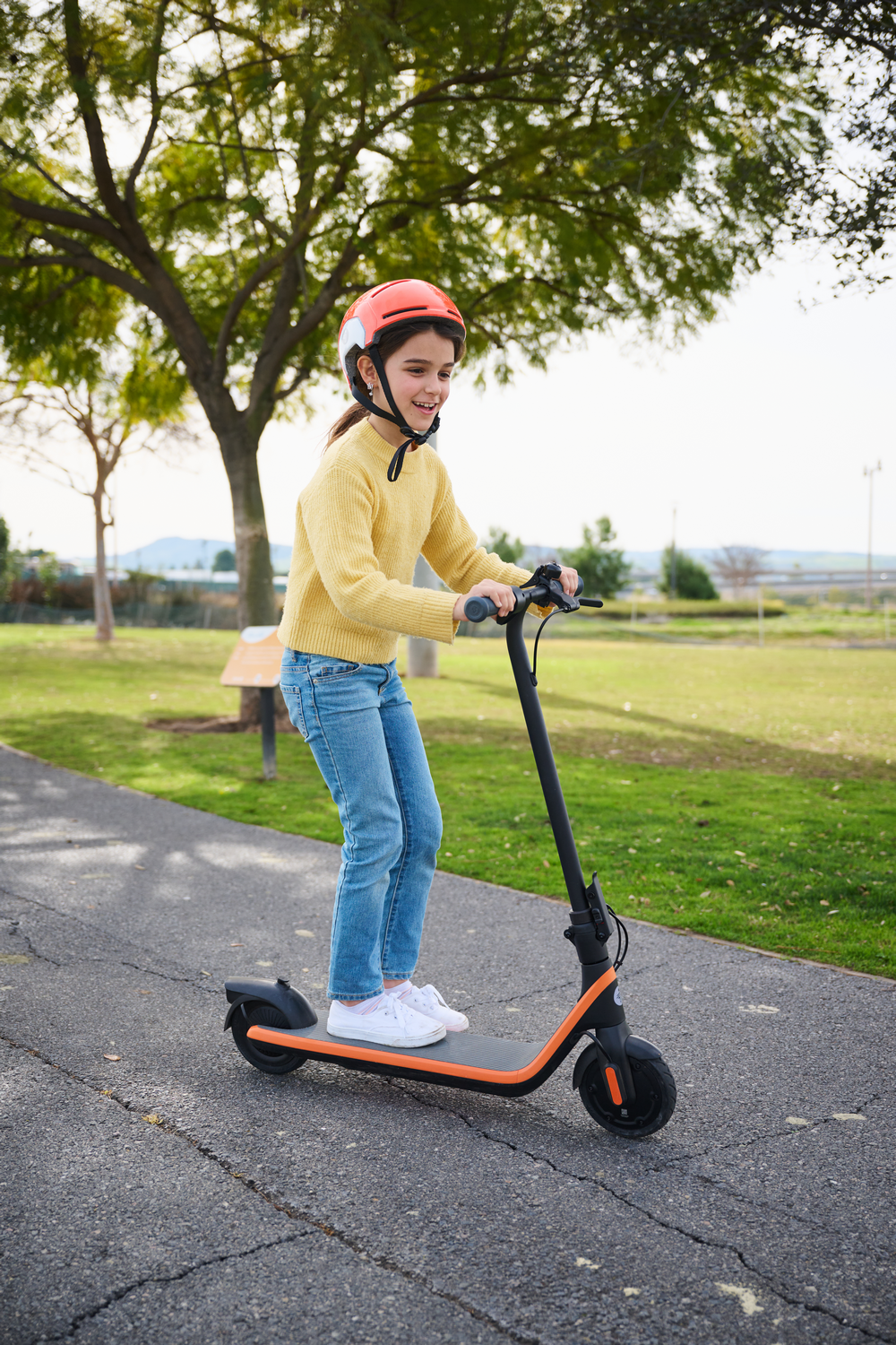 Niña en patinete sonriendo, con casco, en camino pavimentado con césped y árboles al fondo.