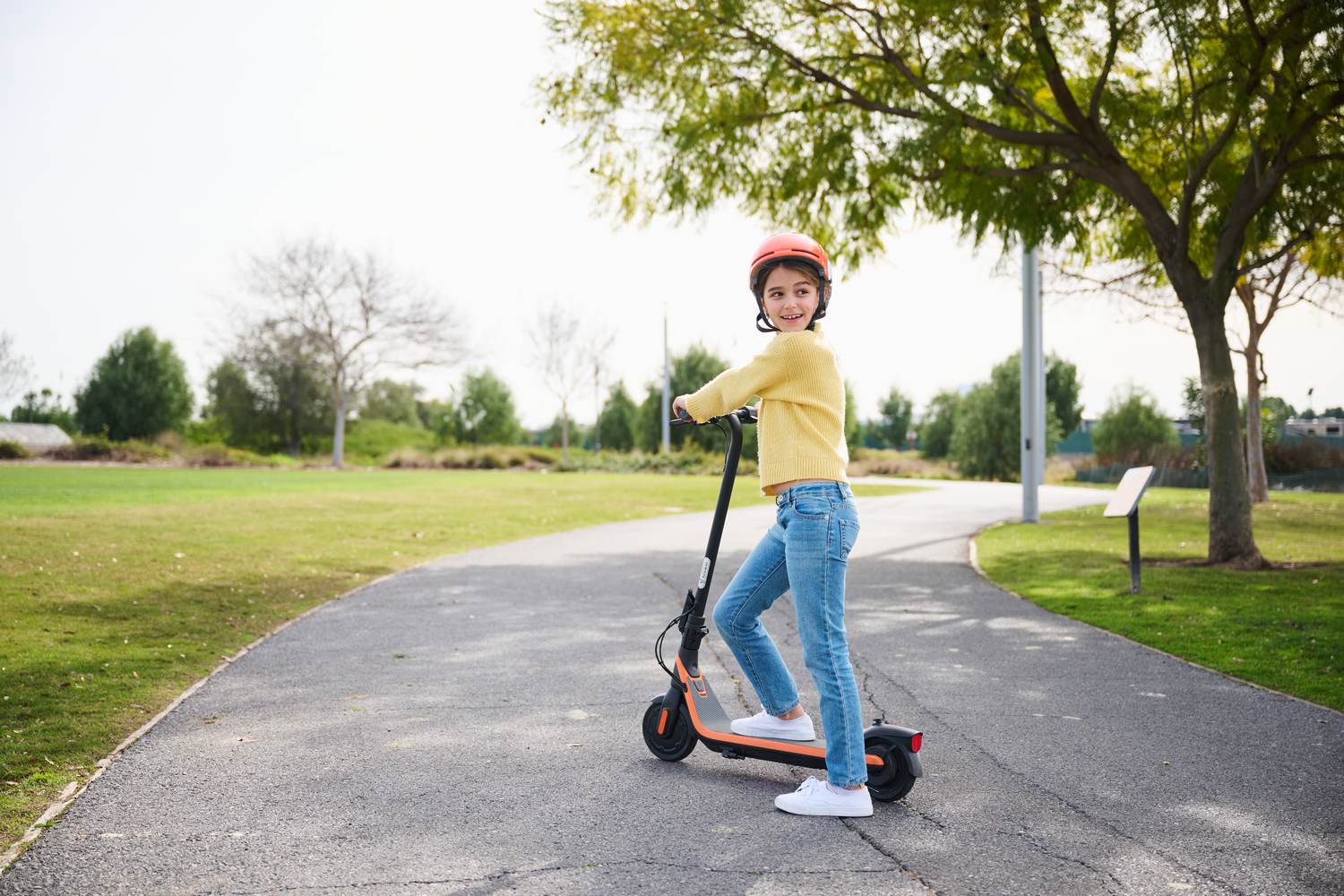 Joven con casco en un scooter en un camino, con un parque verde de fondo.