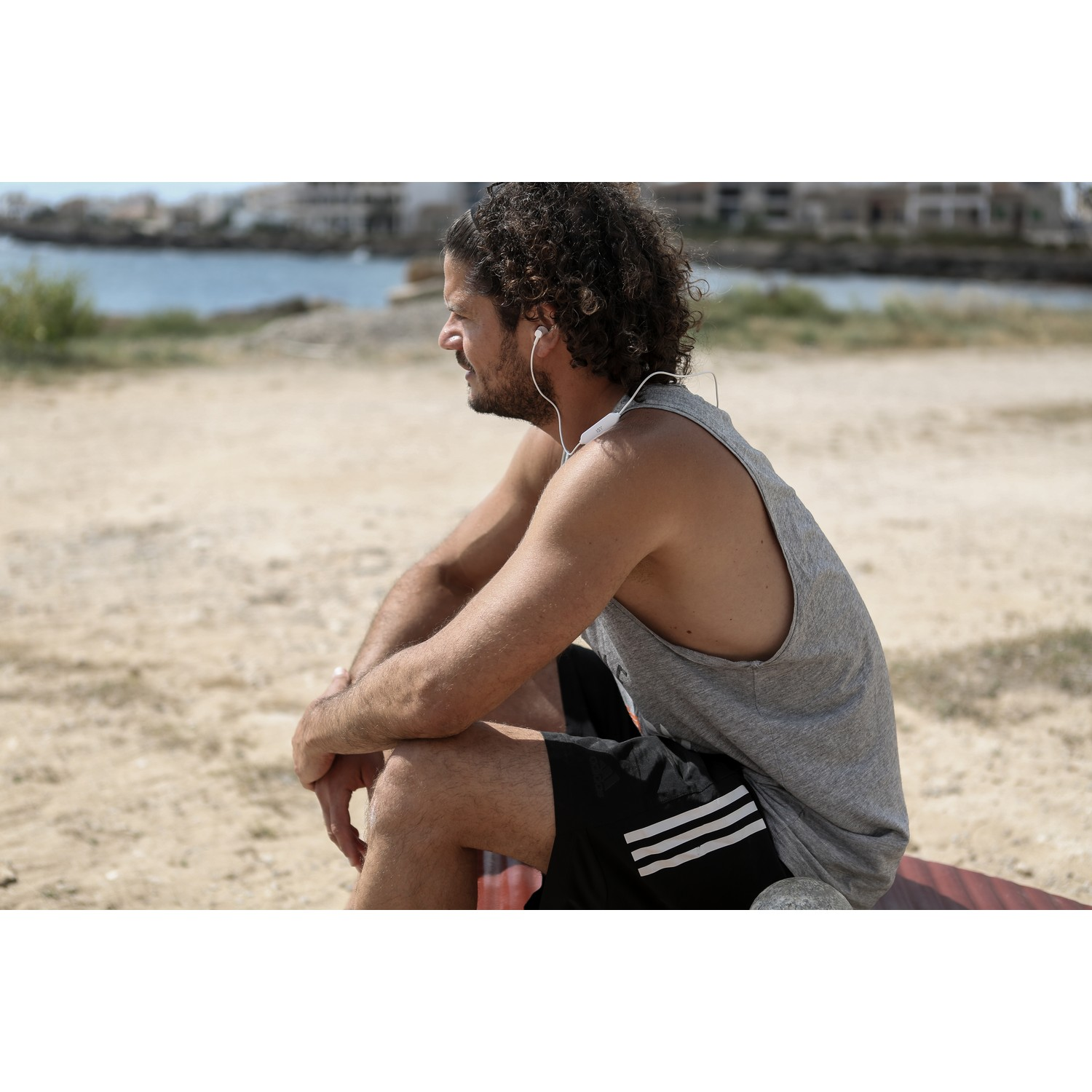 Een man zit op het strand, draagt een grijze tanktop en een zwarte korte broek met witte strepen. Hij draagt oordopjes.