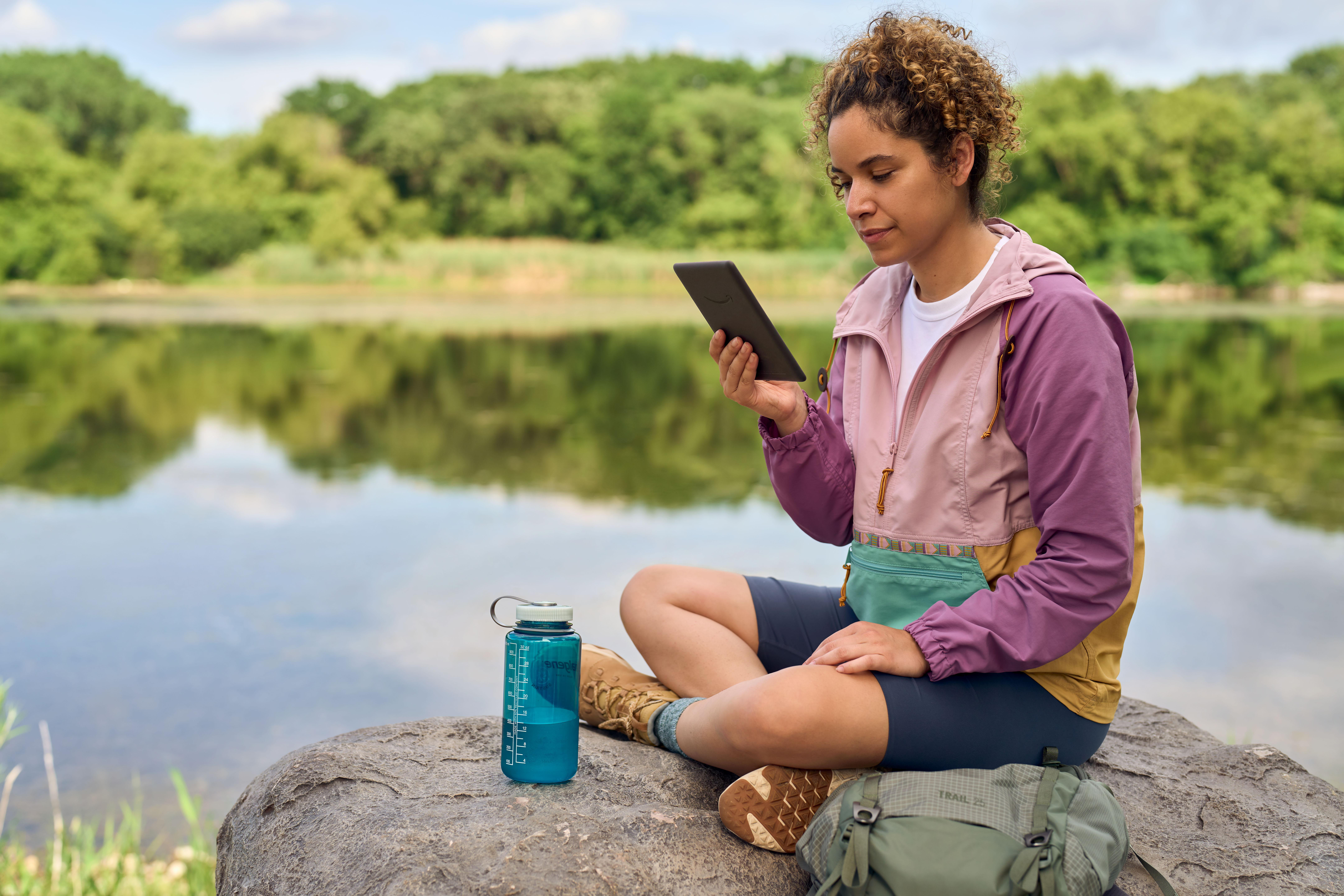 Eine Frau sitzt an einem See und liest ein Tablet, mit einer Wasserflasche und einem Rucksack in der Nähe.