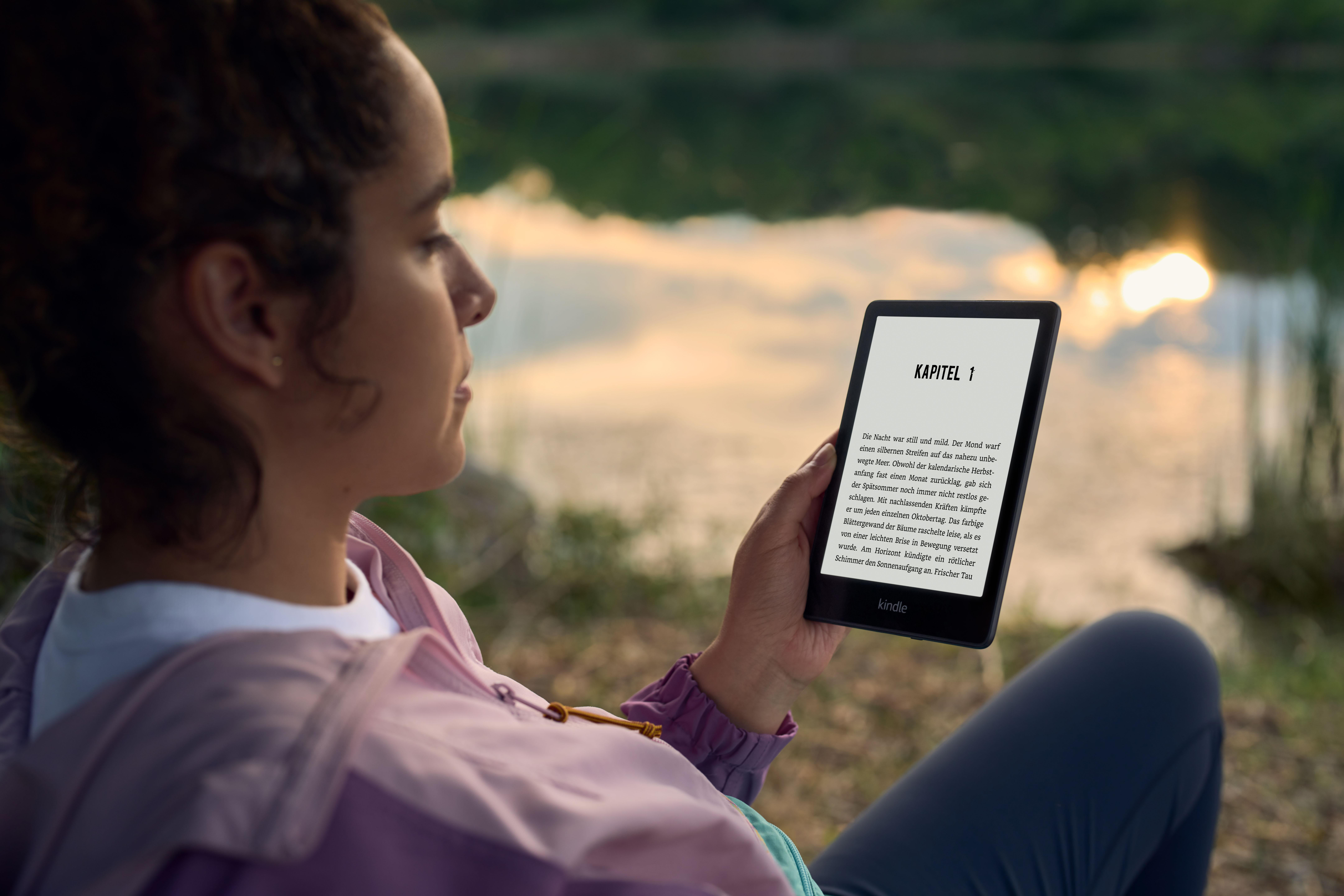 Frau liest einen Kindle-E-Reader im Freien in der Nähe eines Sees. Der Bildschirm zeigt Text.