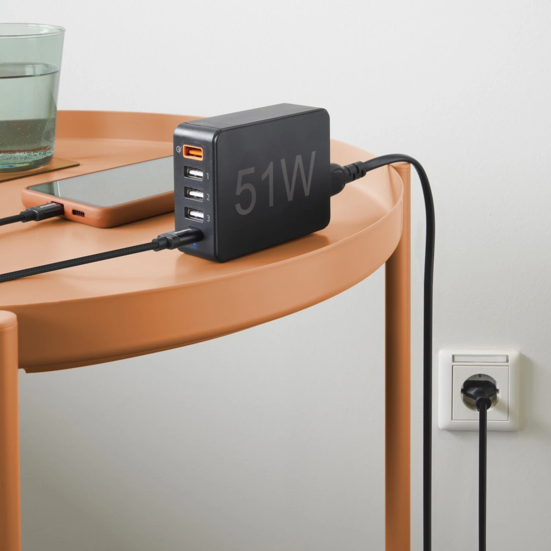 Ein schwarzer Netzadapter mit mehreren Anschlüssen liegt auf einem Tisch, mit Telefon und Wasserglas.