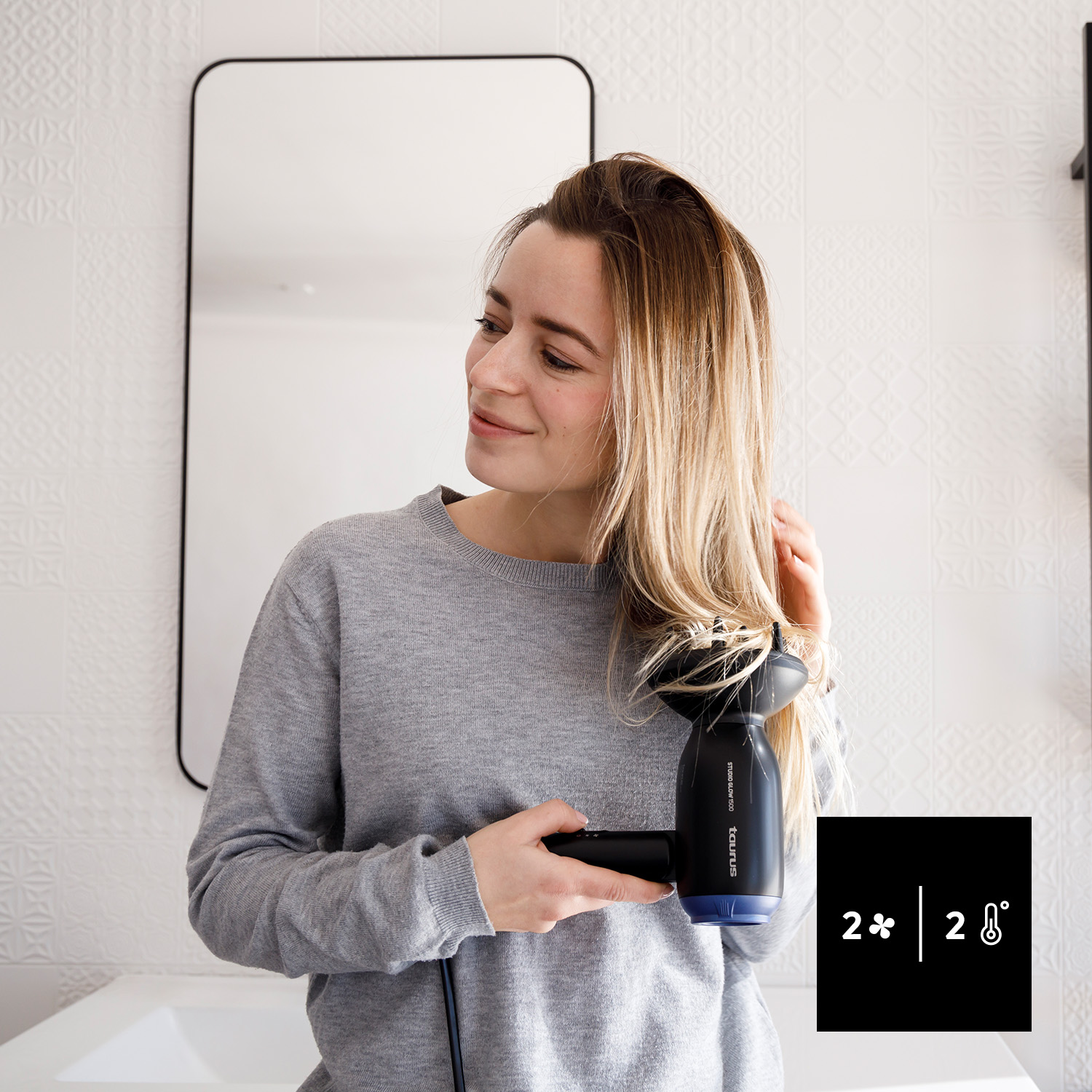 Mujer rubia secando el pelo. Secador, espejo, lavabo, y texto.