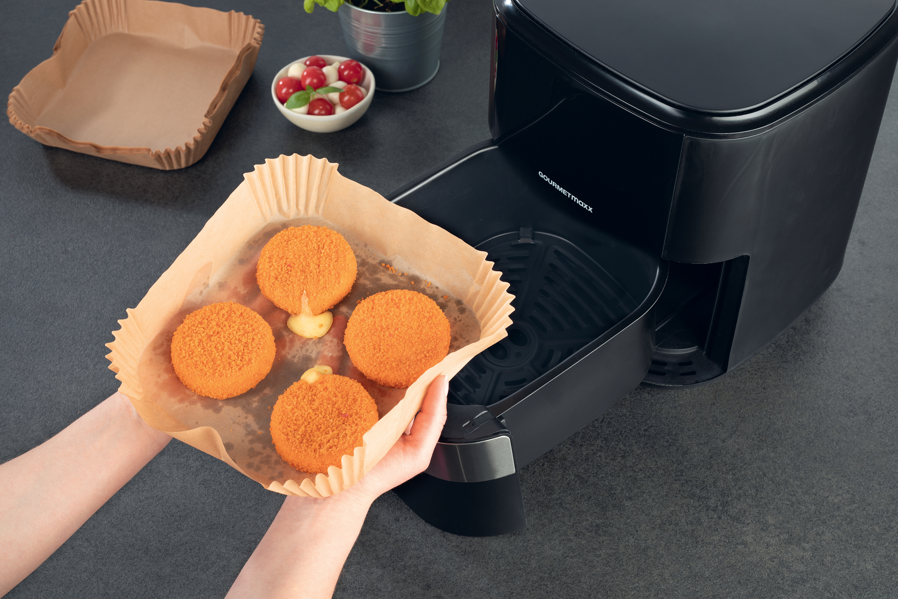 Hände halten Tablett mit frittierten Kroketten, daneben Airfryer auf dunkler Arbeitsplatte.