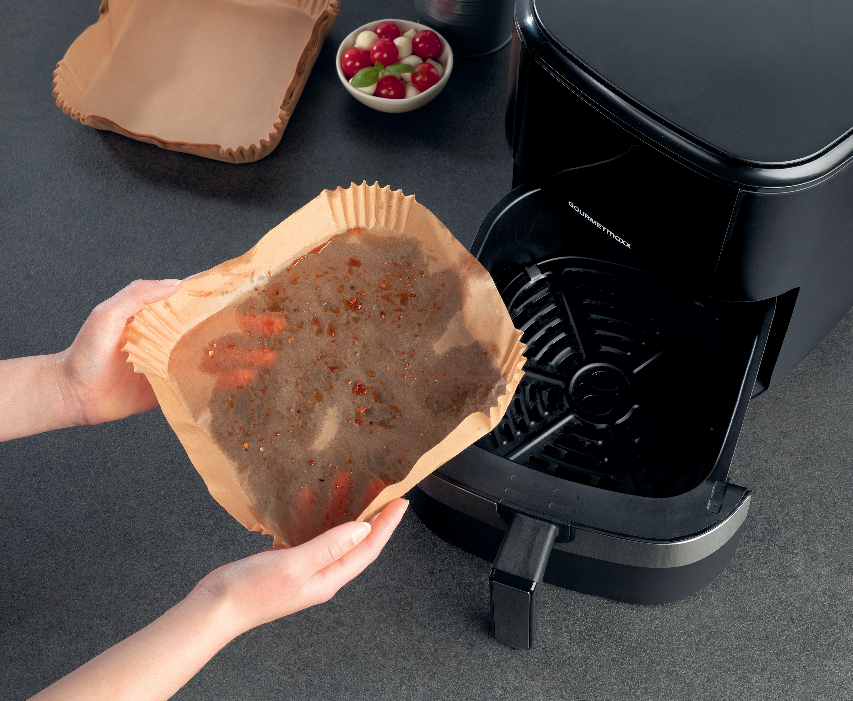 Hände halten ein Tablett mit Essen und legen es in eine schwarze Heißluftfritteuse. Anderes Essen in einer Schüssel.