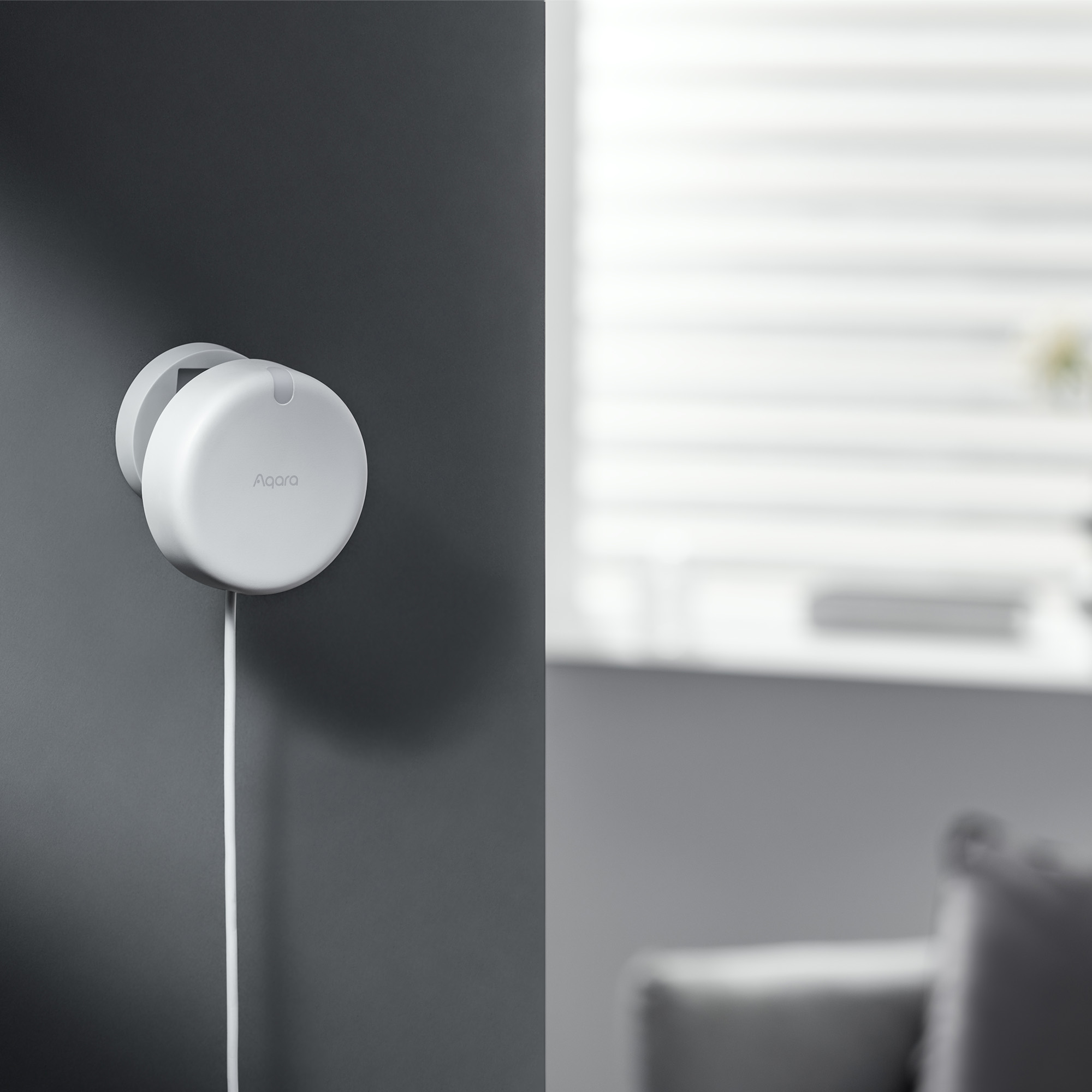 Weißes Aqara Smart Home Gerät an einer grauen Wand montiert. Ein weißes Kabel hängt von dem Gerät.