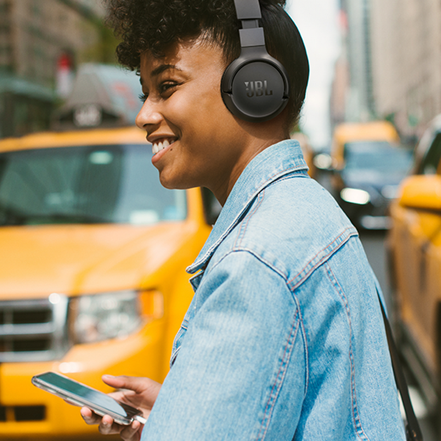 Smiling woman with JBL headphones on a city street, holding a phone, yellow taxi in background.