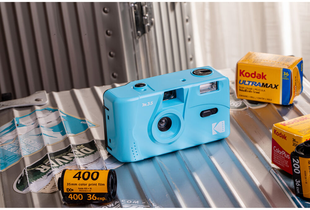 Világoskék Kodak kamera filmekkel és vakuval, ezüst bőröndön matricákkal.