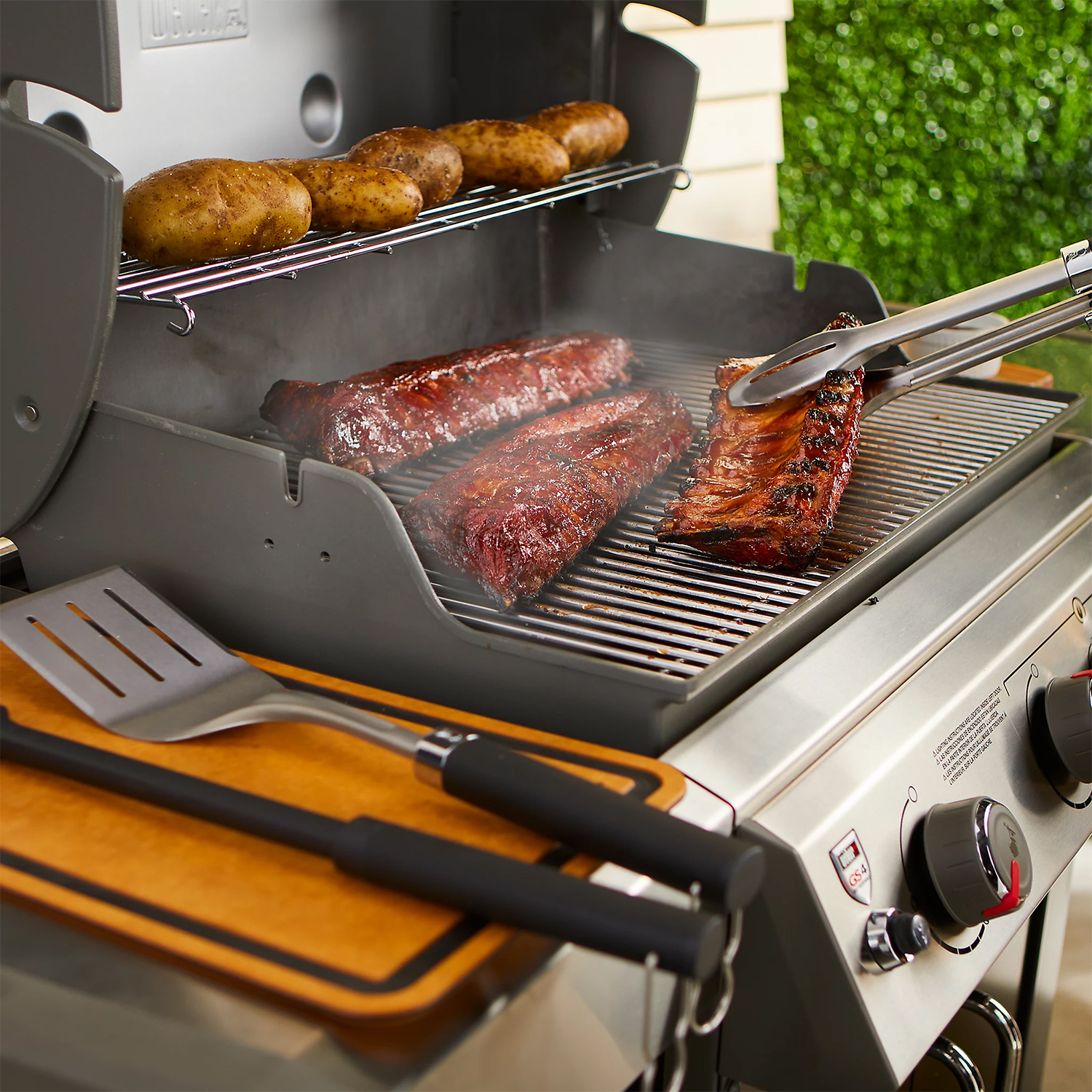 Un grill con costine e patate in cottura. Pinze e una spatola a lato.