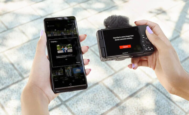 Dłonie trzymające telefon i aparat, przesyłające obrazy. Telefon pokazuje pasek postępu, aparat 'importowanie'.